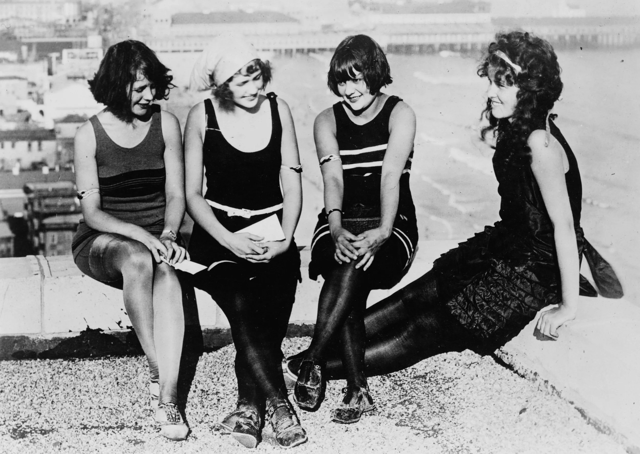 Four young women sitting together on a ledge outdoors with a cityscape in the background, smiling and talking.