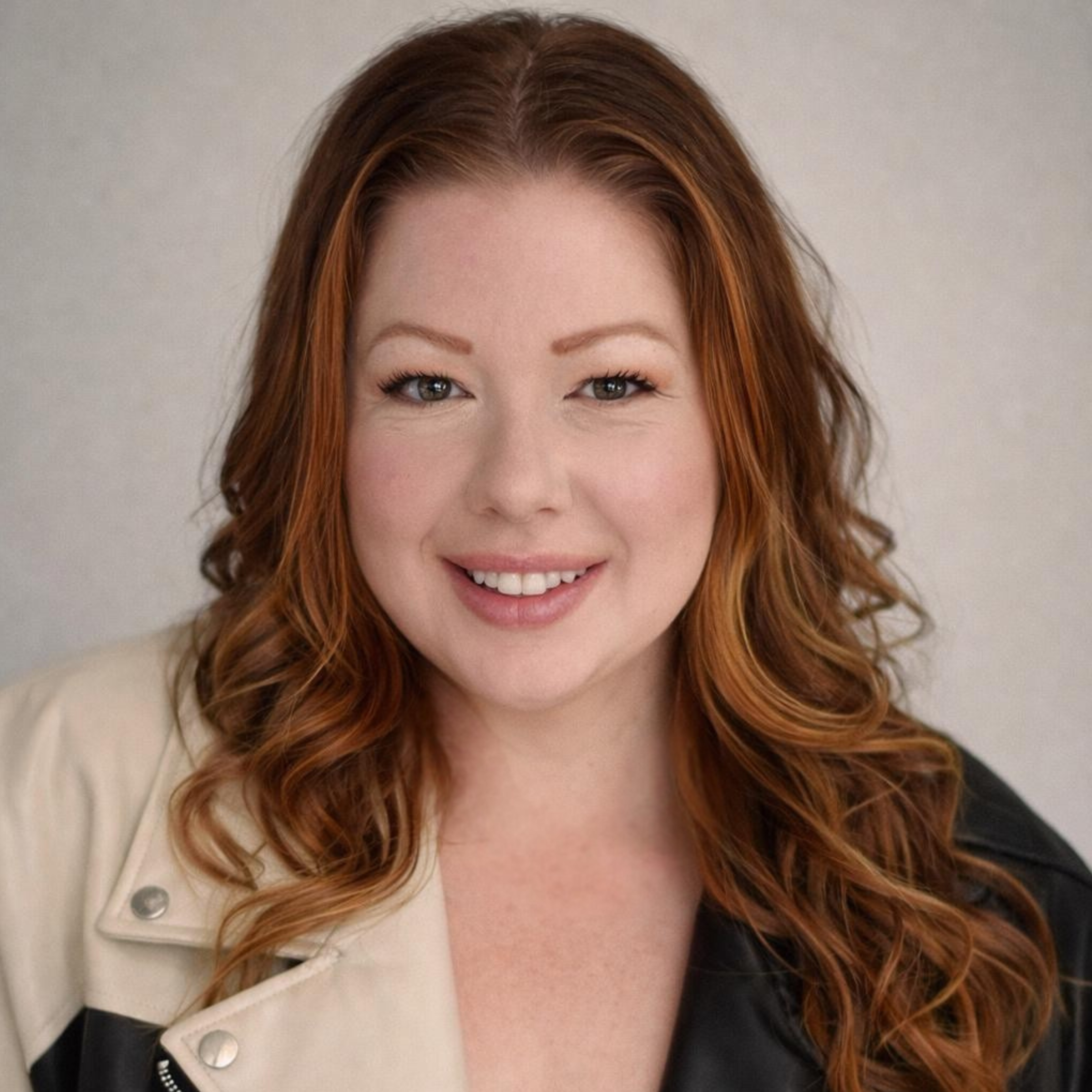 Close-up portrait of a smiling woman with red hair, wearing a beige and black jacket, against a light neutral background.