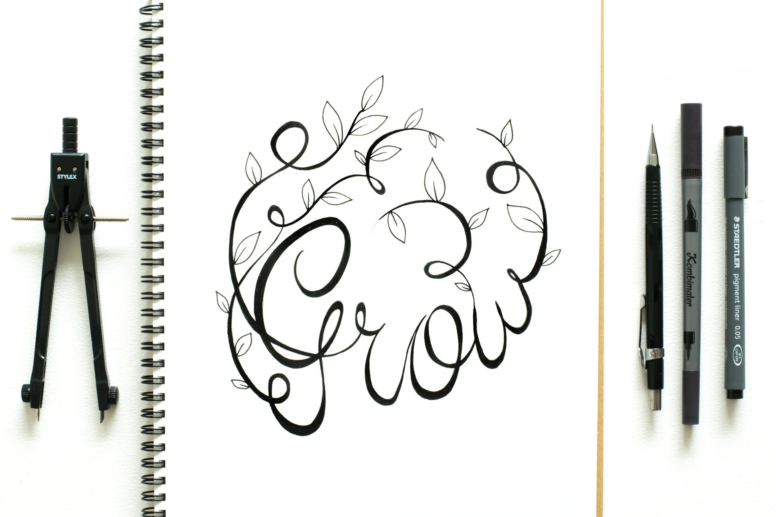 A blank sketchbook with a black ink drawing of the words 'Thank You' in cursive, surrounded by leaves and vines, with drawing tools (compass, mechanical pencil, gel pen, marker) laid out on a white surface.