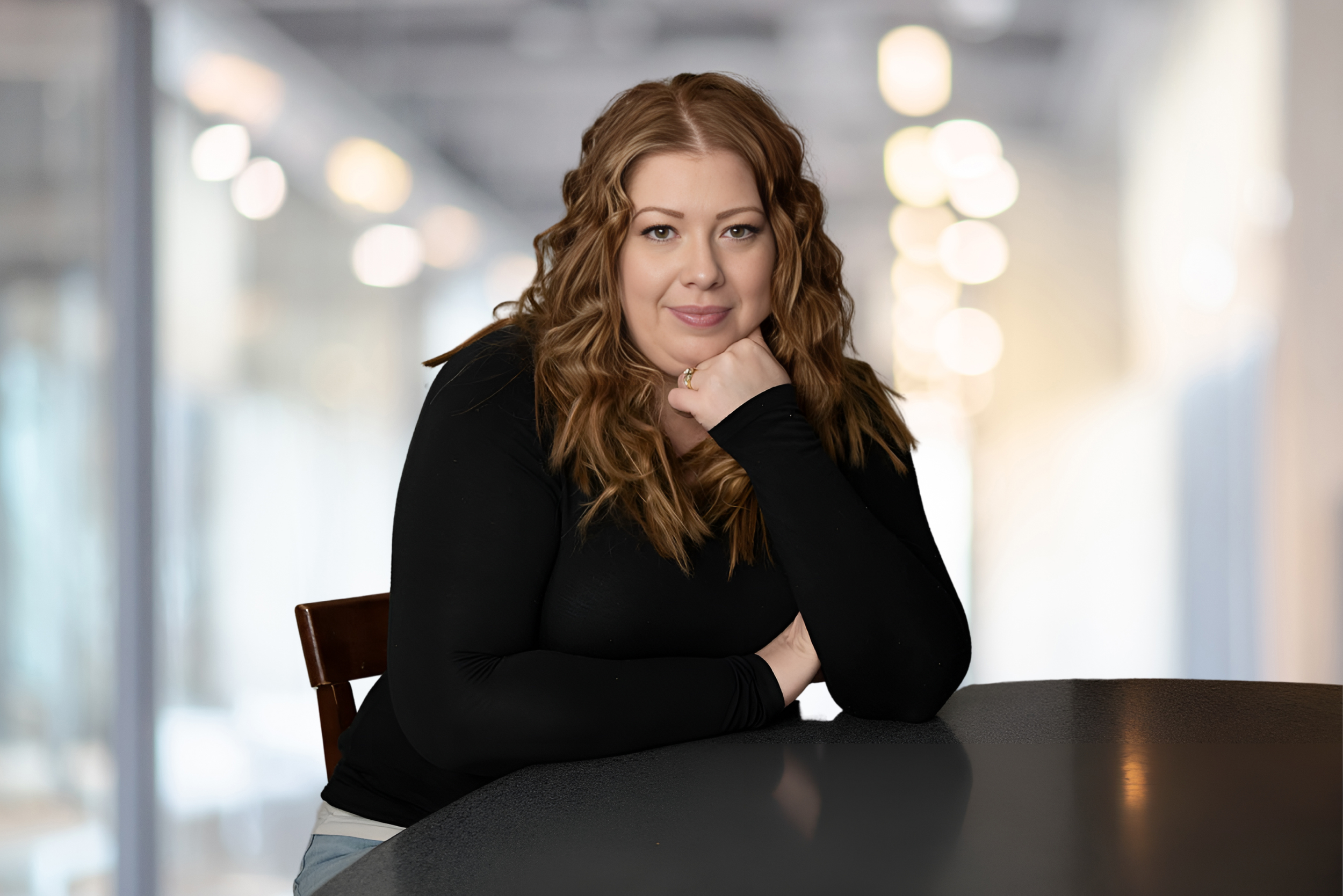A woman with long, curly red hair, fair skin, and wearing a black long-sleeve top, sitting at a dark table with a blurred indoor background featuring bokeh lights, resting her chin on her hand and smiling softly at the camera.