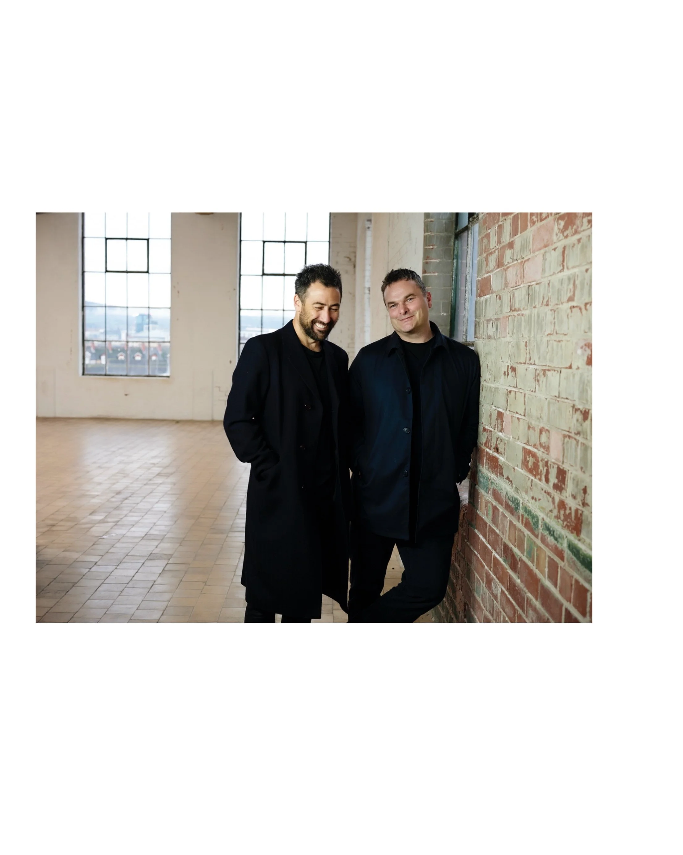 image of two men leaning against brick wall. wearing black