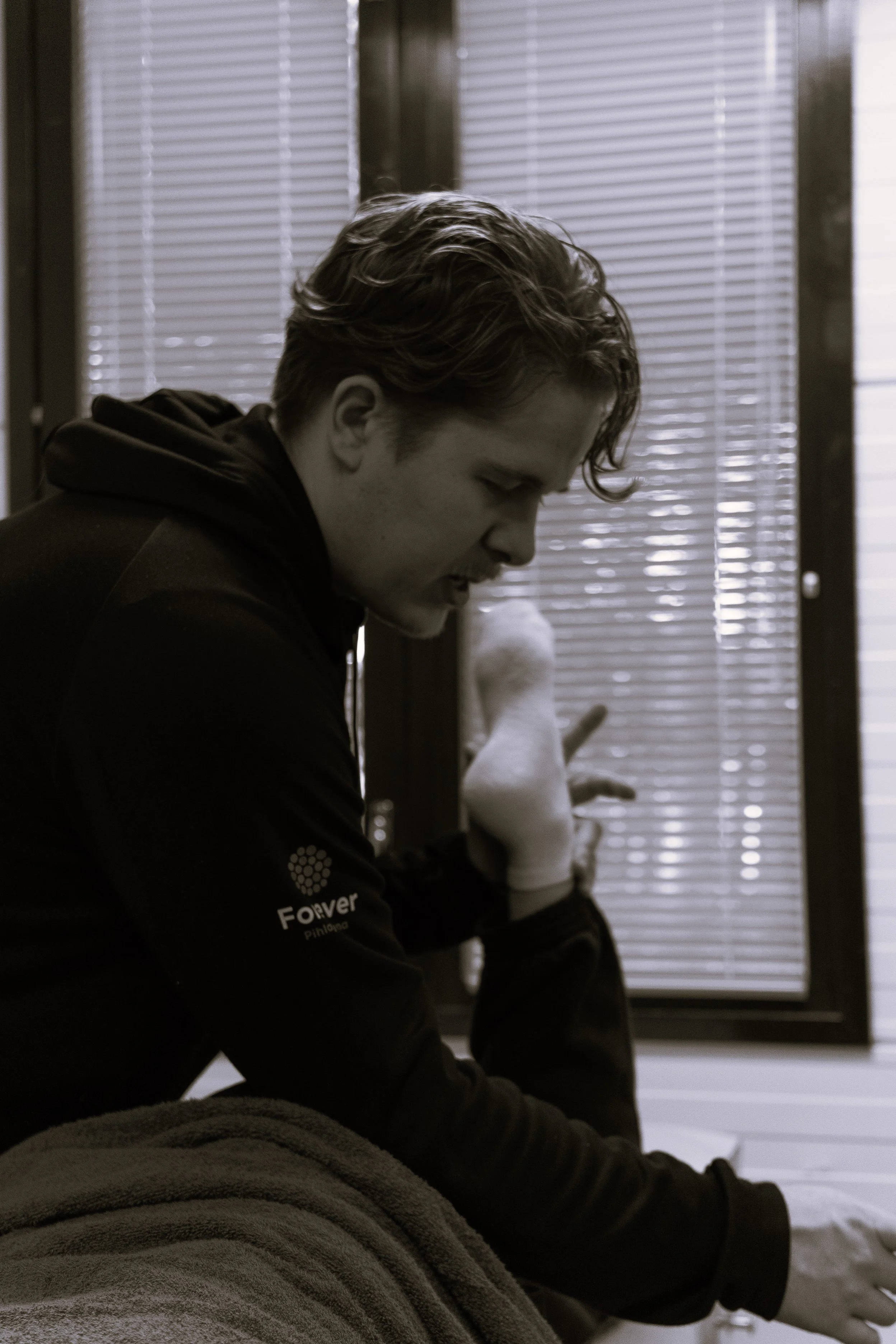 A young man with wavy hair wearing a hoodie sits next to a window with blinds, looking down with a focused expression.