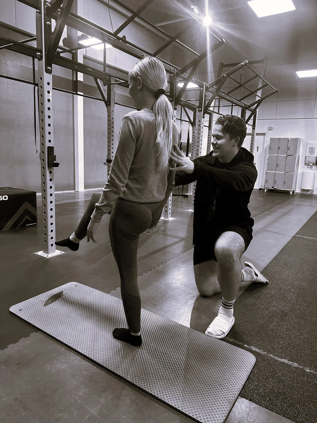 A woman with long blonde hair in athletic clothing is standing on a mat in a gym while a man kneels beside her, giving her a shoulder massage.