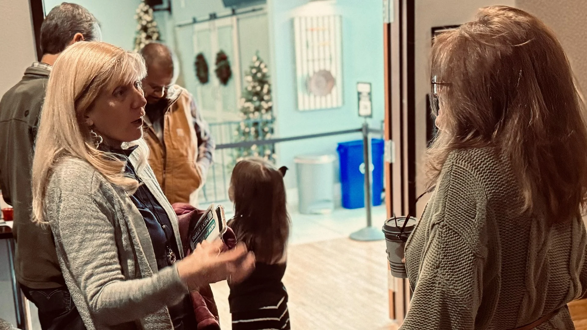 Three women and a young girl are in conversation inside a building decorated with Christmas wreaths and a Christmas tree, with a blue recycling bin and a blue trash can in the background.