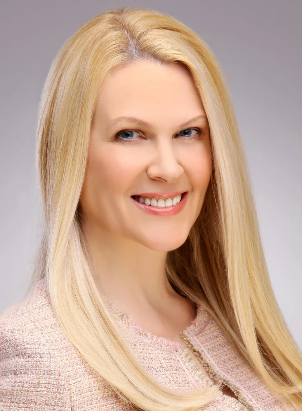 Smiling blonde woman with long straight hair wearing a pink textured jacket against a gray background