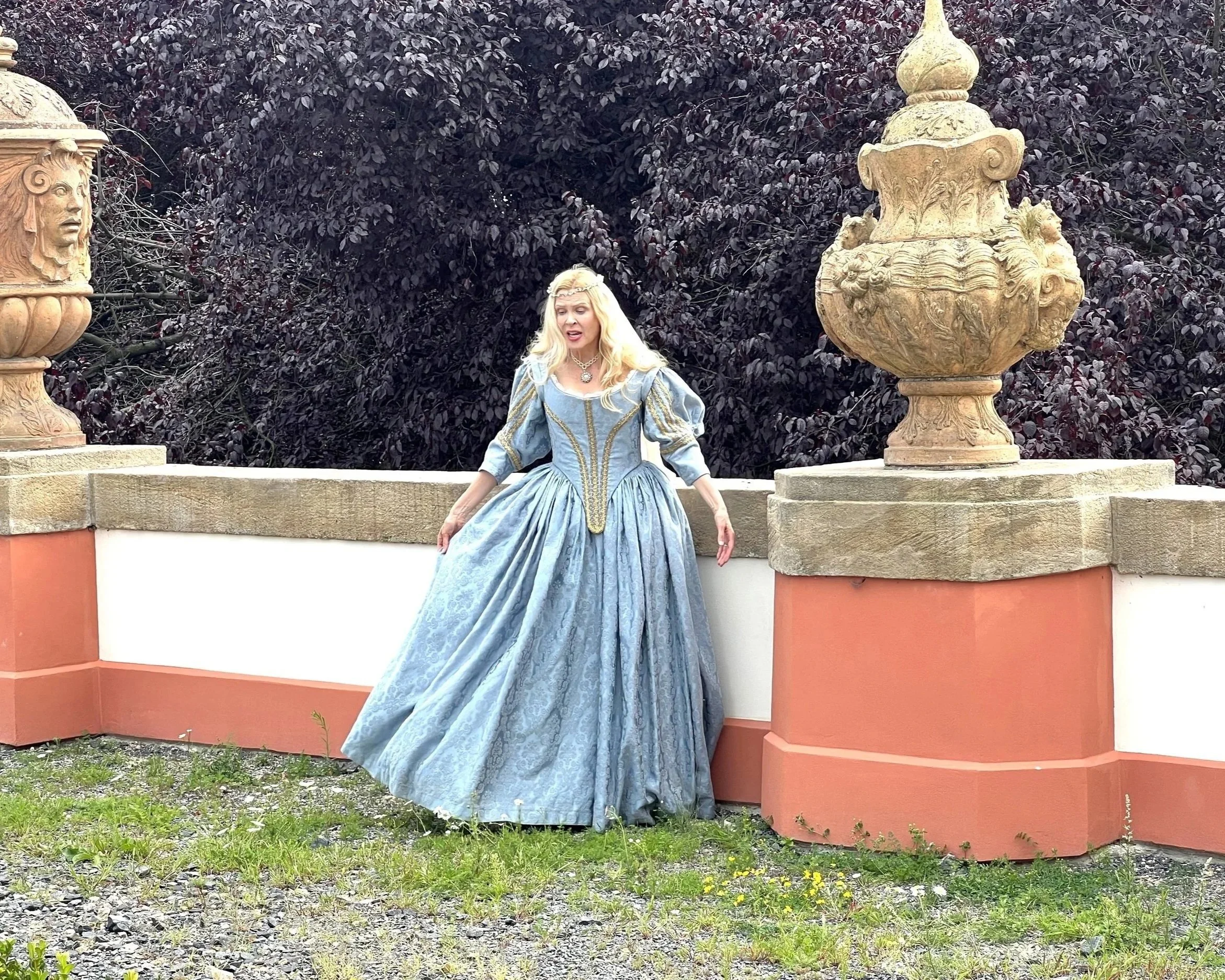 A person dressed in 17th century historical aristocratic costume in a blue gown with gold embroidery, standing outdoors in historical European garden between two ornate stone fountains, with dark purple foliage in the background.