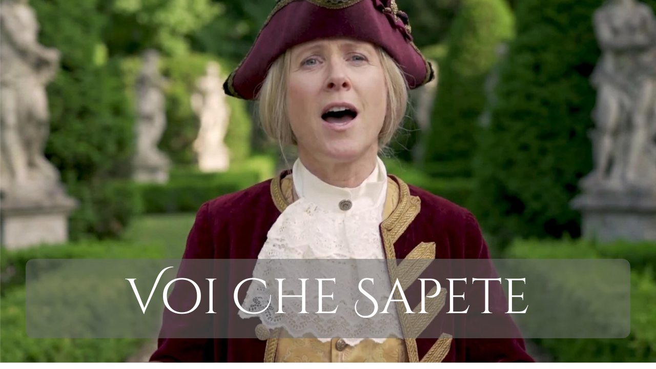 A woman dressed in historical clothing, wearing a burgundy jacket and a tricorn hat, standing outdoors with green trees and statues in the background, singing or speaking with her mouth open. The text "VOI CHE SAPETE" appears across the bottom of the image.
