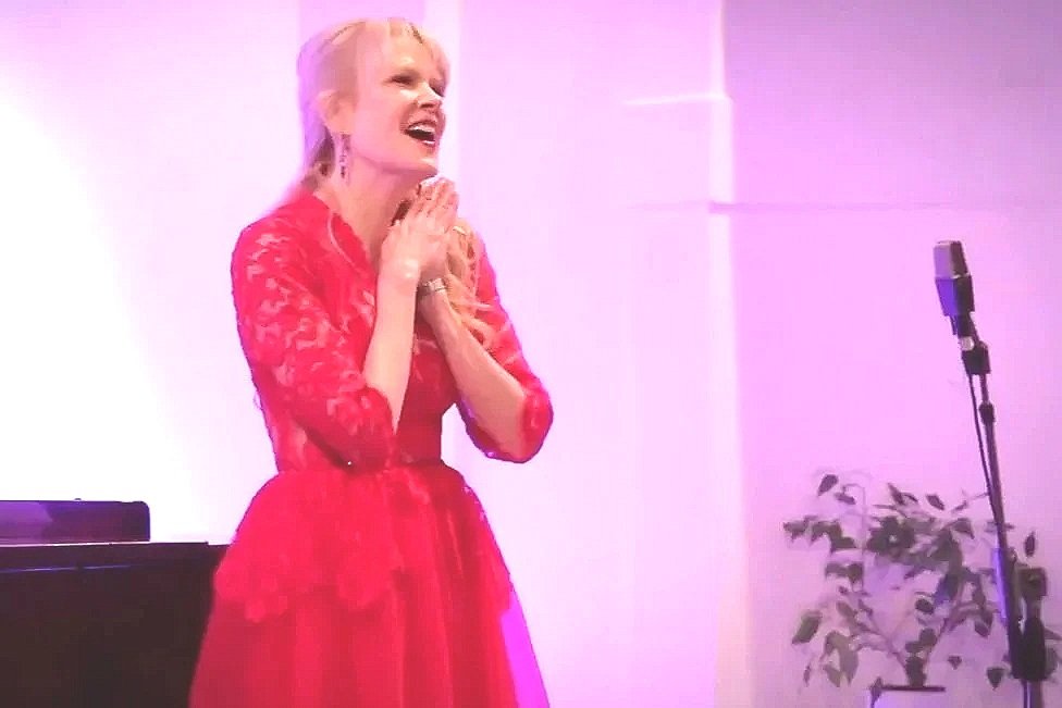 A woman in a red lace dress standing in front of a microphone with a piano and a plant nearby, smiling and clasping her hands together.
