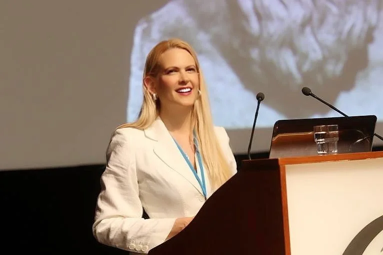 A woman with long blonde hair, wearing a white blazer and a conference badge, standing at a podium and smiling during a presentation or speech.