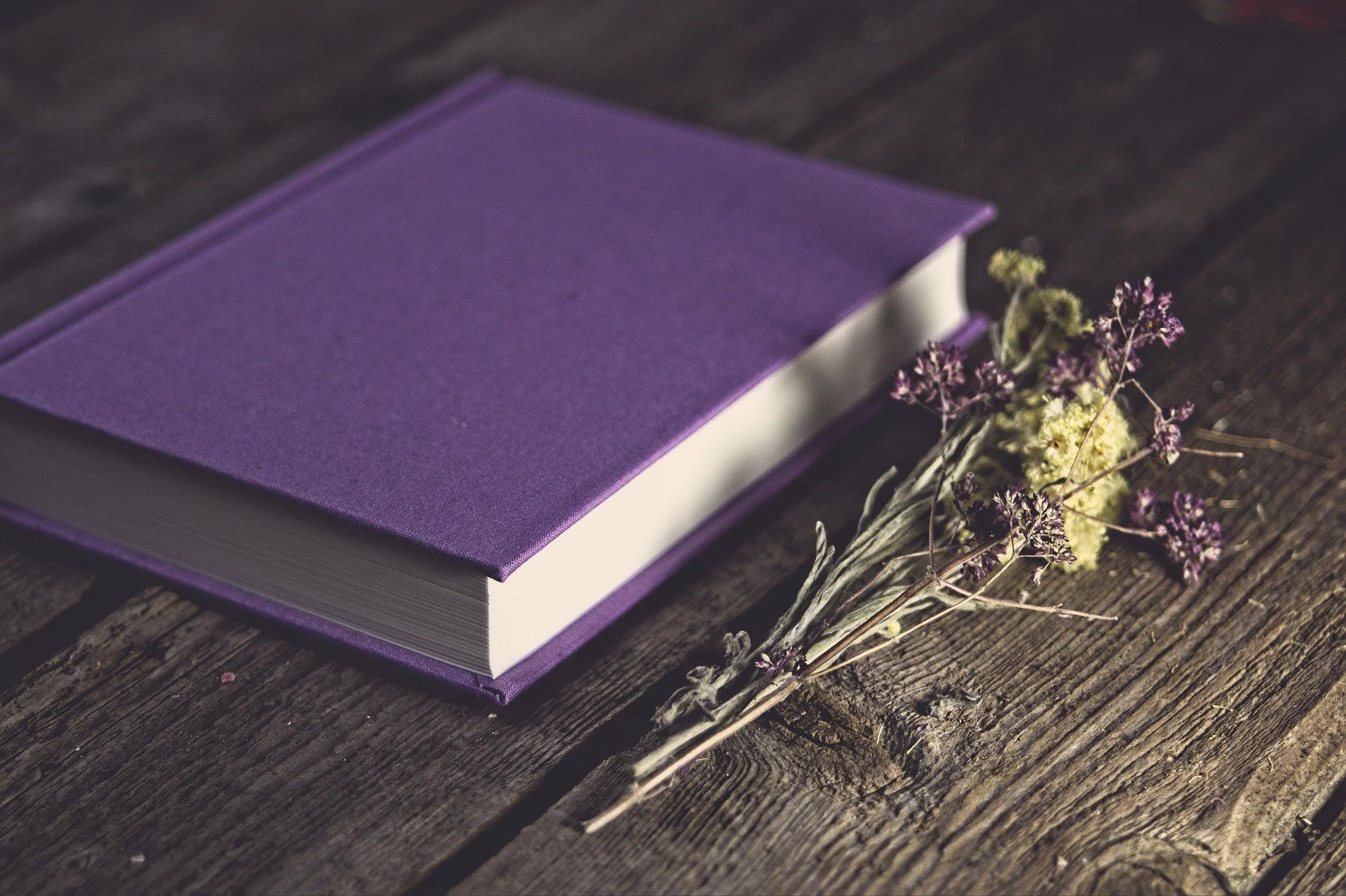 A closed purple hardcover book resting on a wooden surface next to dried purple and yellow flowers.