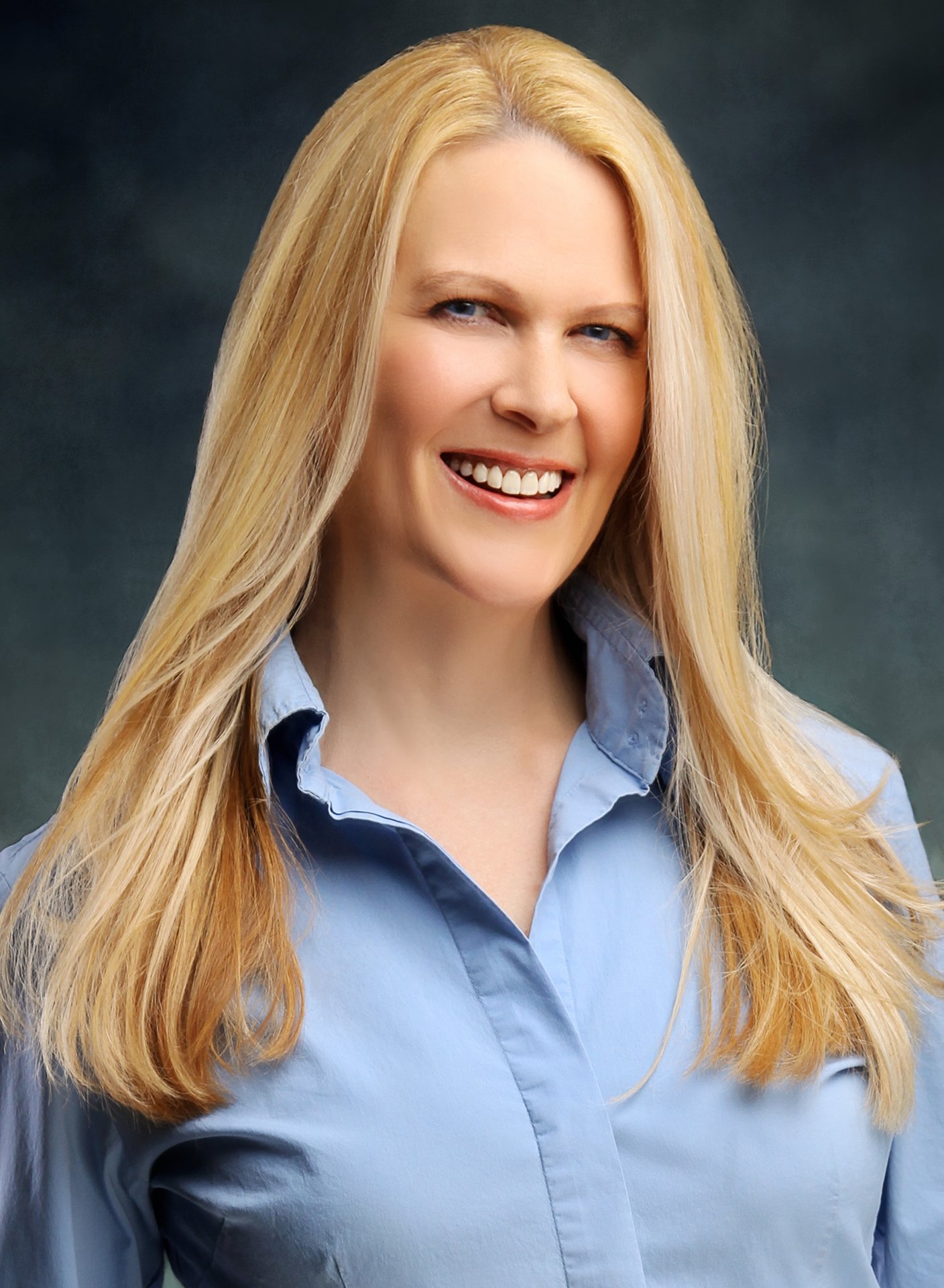 Head and shoulders portrait of a smiling woman with long blonde hair, wearing a light blue collared shirt against a dark background.