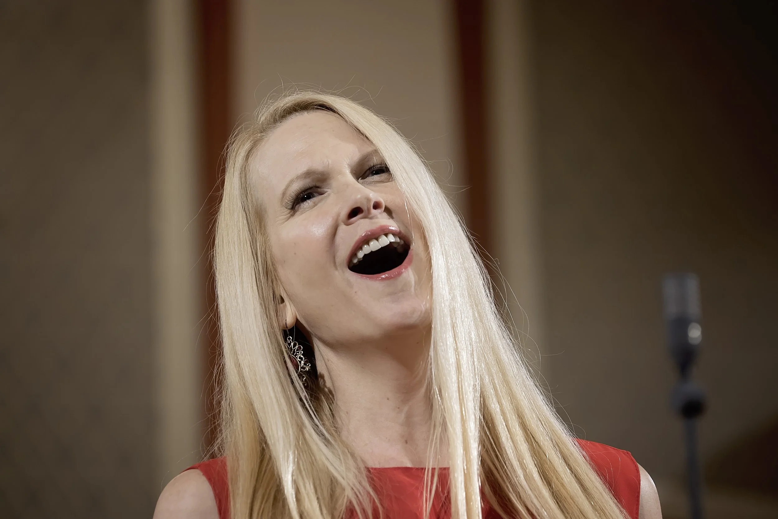 A smiling blonde woman wearing a red dress with long hair, earrings, and makeup, singing or speaking into a microphone in Suk Hall, Rudolfinum.