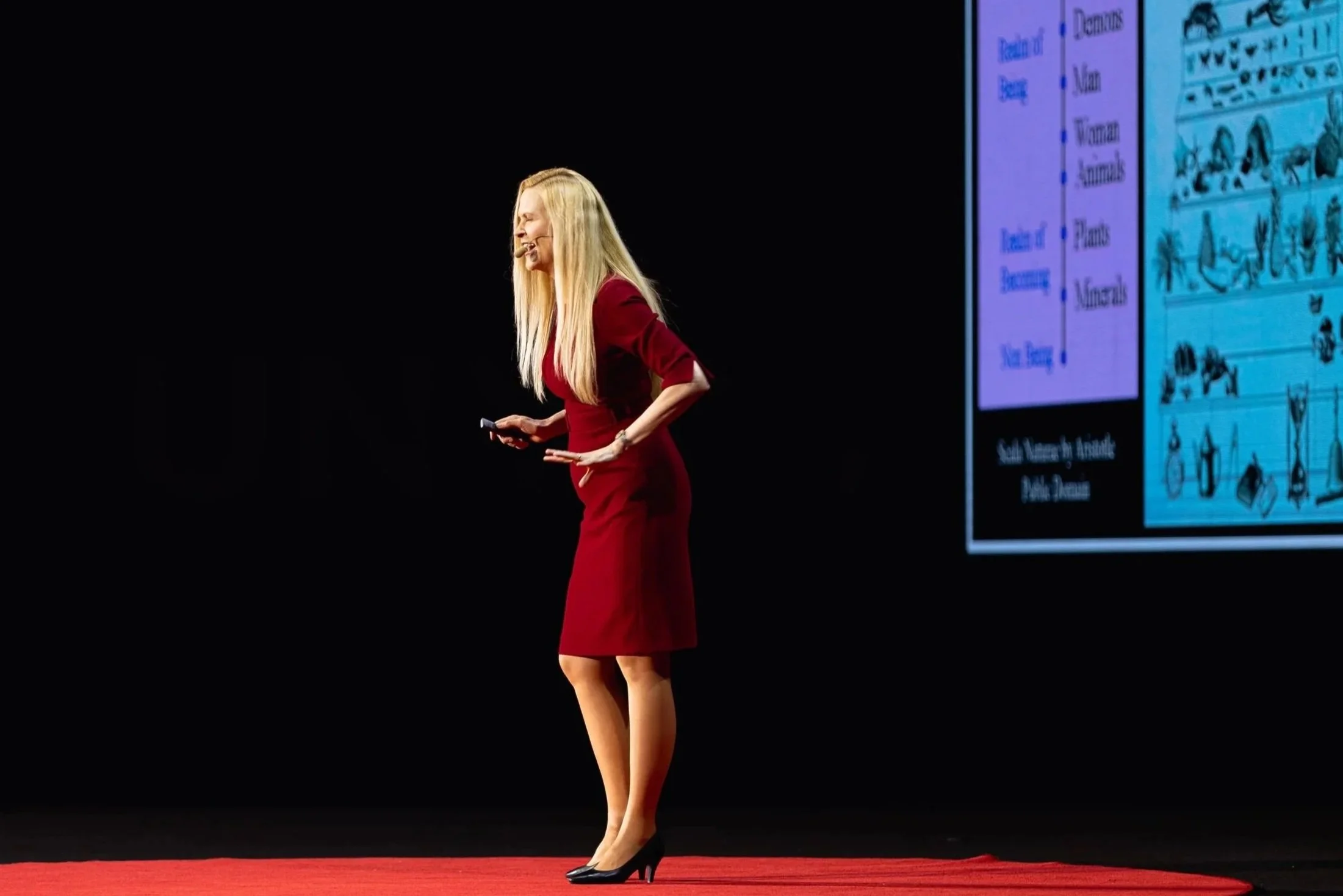 A woman with long blonde hair in a red dress and black high heels standing on a red carpet, smiling and speaking into a microphone headset, with a presentation slide behind her.