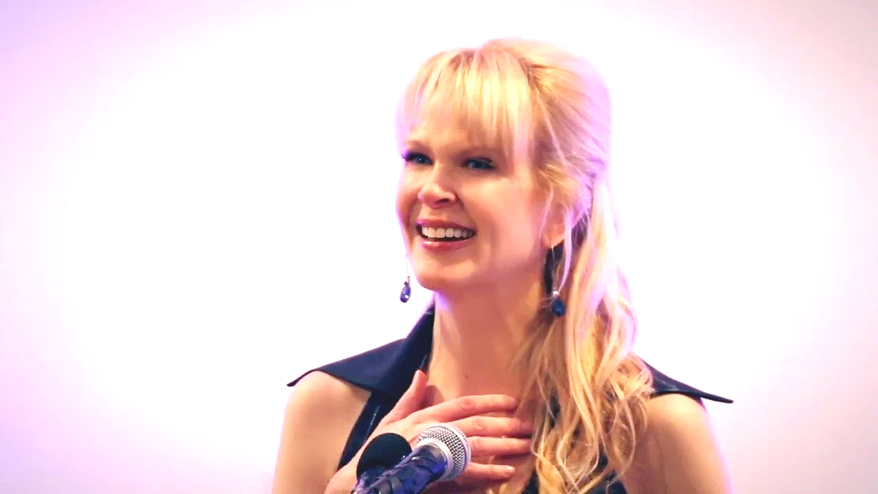 A woman with blonde hair and black earrings smiling and holding her hand to her chest.