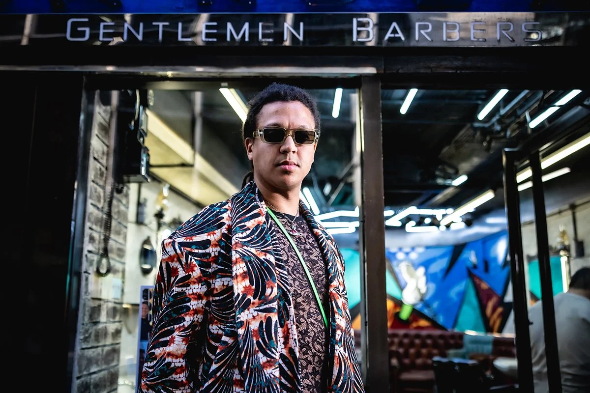 A man wearing dark sunglasses and a colorful, patterned blazer standing in front of a barbershop sign that says 'Gentlemen Barbers'.