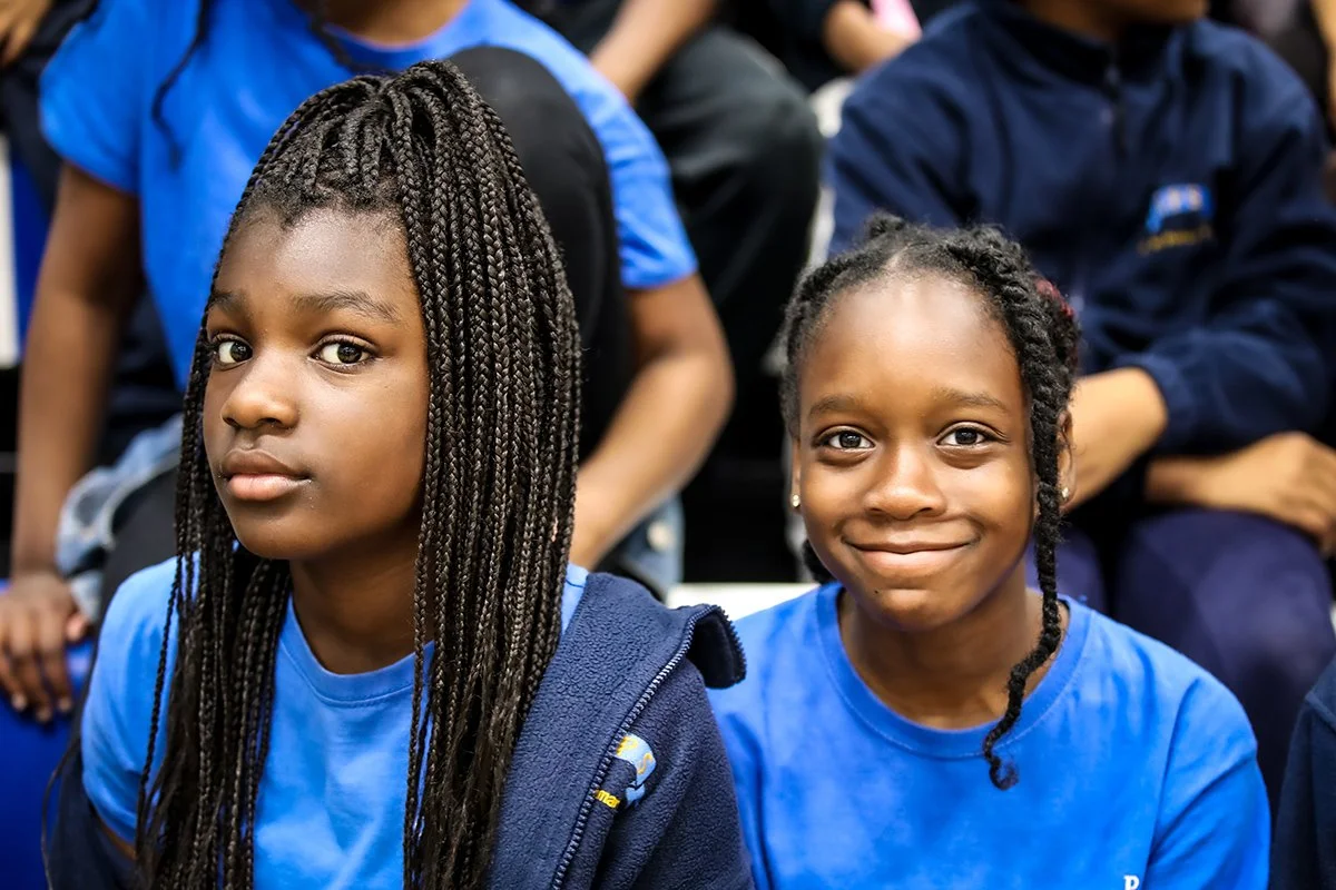 Two young girls sitting in a group, wearing blue shirts, one with long braids and the other with short twisted hair, in an indoor sporting event.