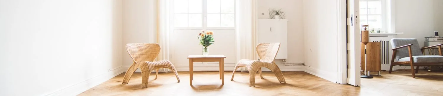 Ein Wohnzimmer mit zwei Korbstühlen, einem kleinen Tisch mit Blumen und einem großen Fenster im Hintergrund.