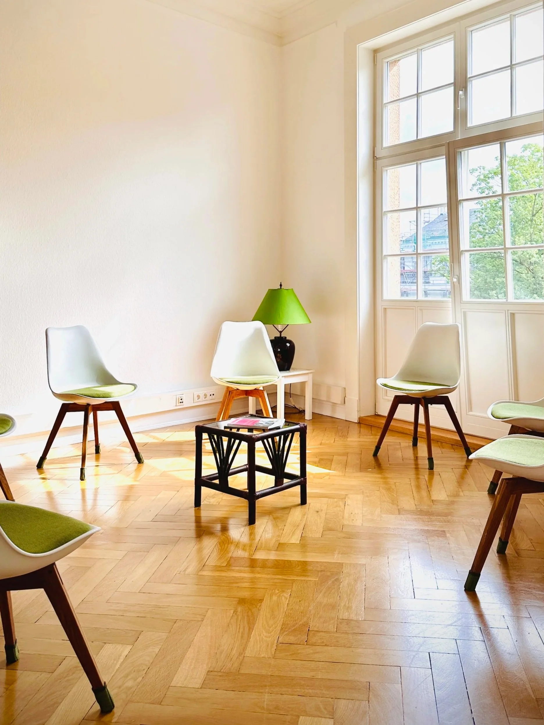 Ein heller, leeres Zimmer mit Holzboden, einem großen Fenster, einem grünen Lampenschirm, mehreren weißen Stühlen mit grünen Kissen und einem kleinen schwarzen Beistelltisch.