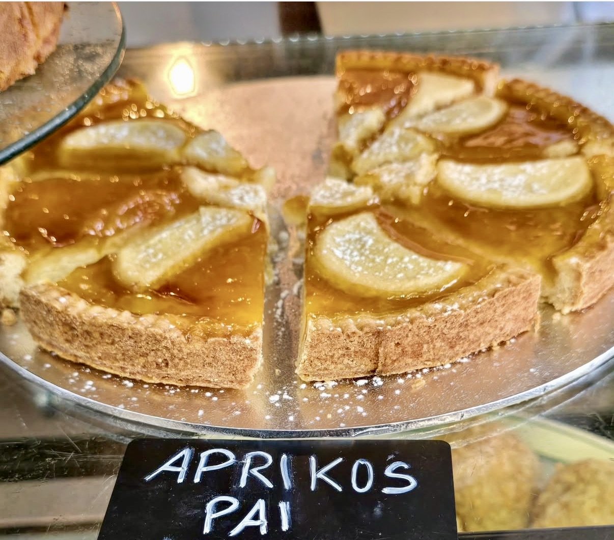 A sliced apricot tart with a golden crust, topped with fresh apricot slices and a shiny glaze, displayed on a glass platter at a bakery or market.