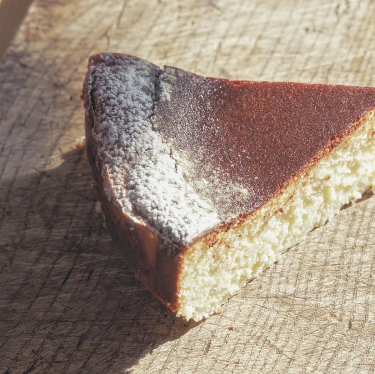 A slice of cheesecake with a chocolate crust on a wooden cutting board, dusted with powdered sugar.