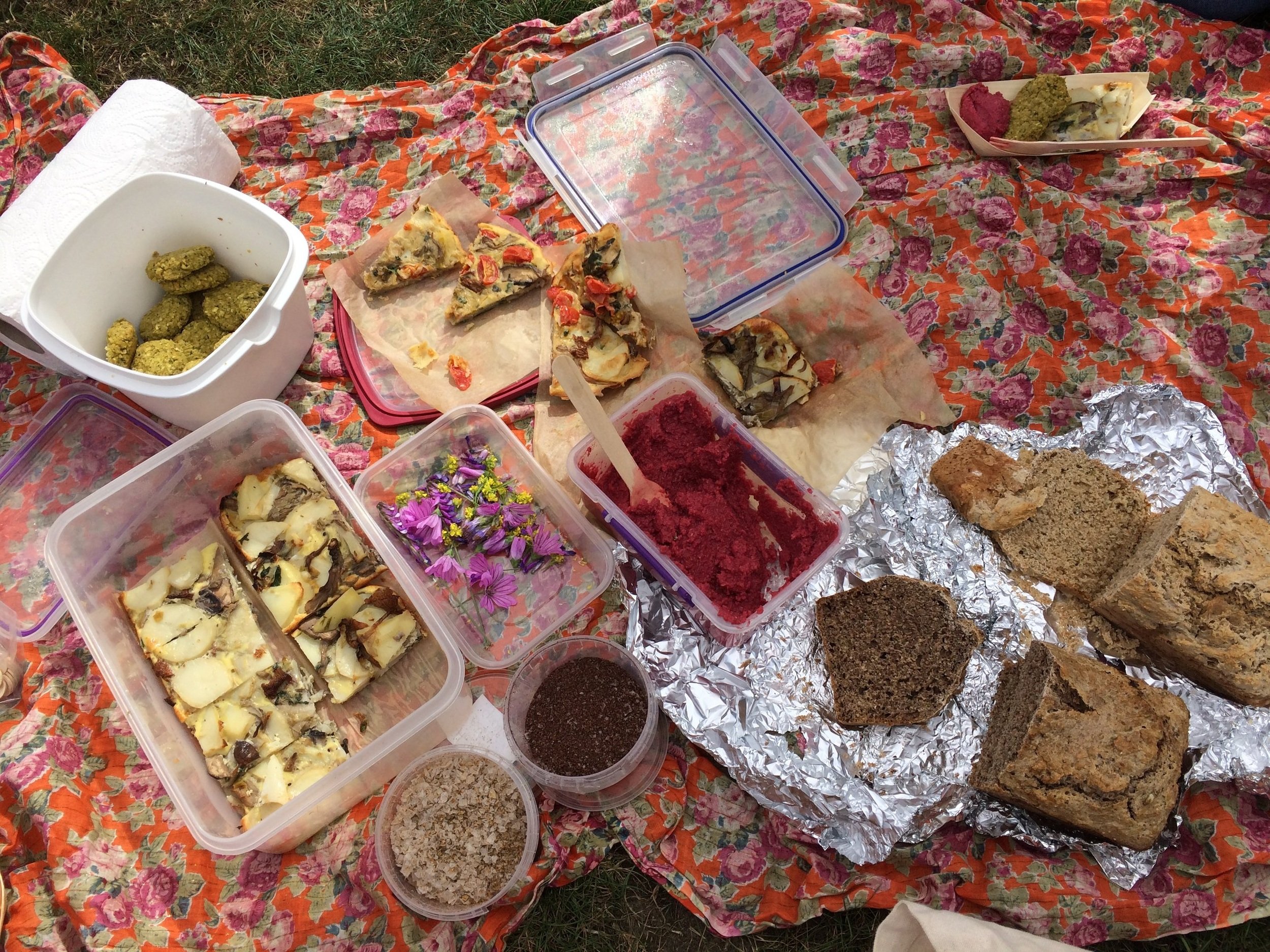 Picnic spread on a floral blanket, featuring homemade bread, beet dip, sliced frittata with vegetables, green cookies, and edible flowers in containers.
