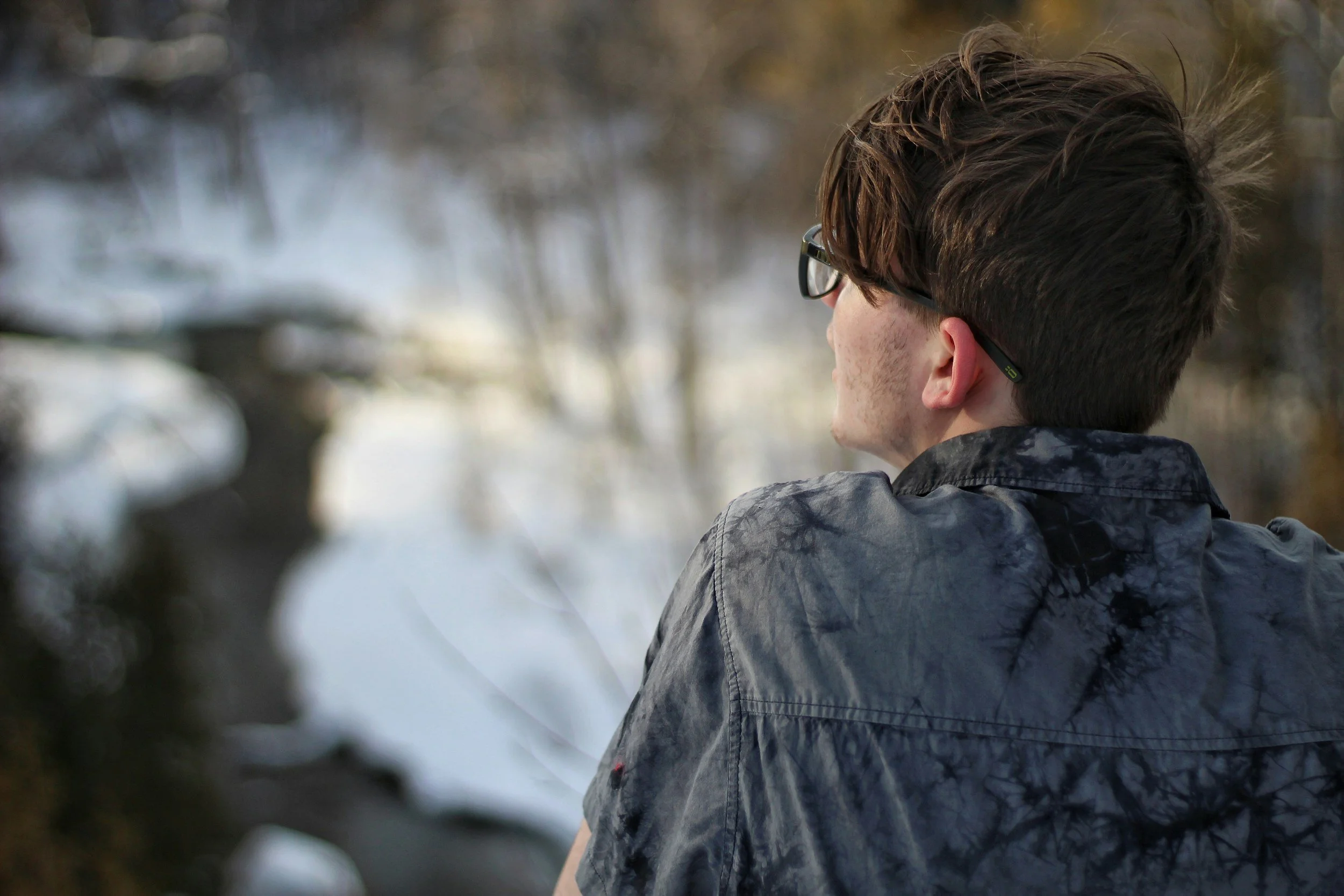 Young adult male with glasses and wearing a blue shirt, sitting near water where trees are reflected.