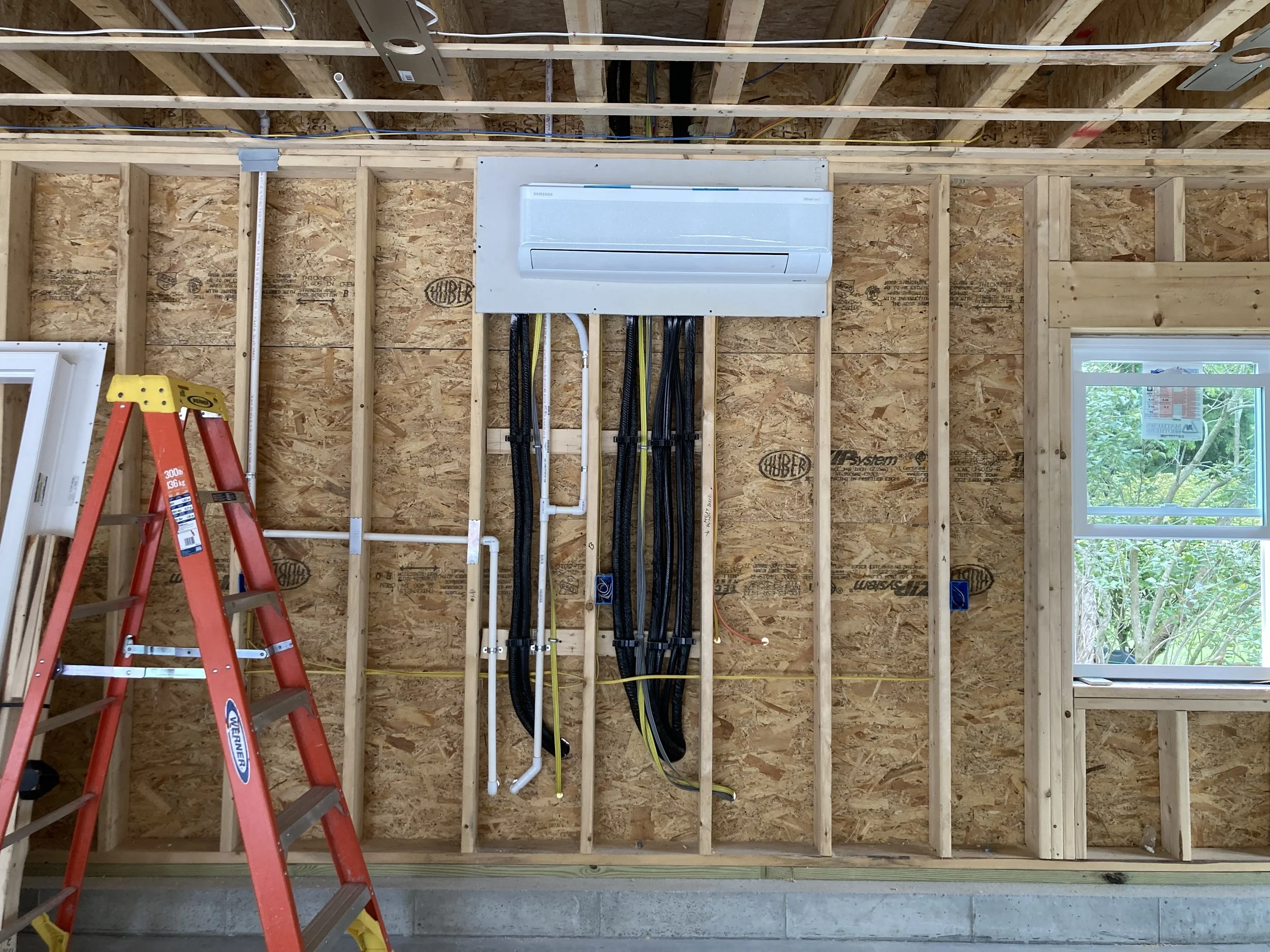 Interior of a building under construction with exposed wooden framing, electrical and plumbing conduit, an air conditioning unit, a red ladder, and a window showing outside trees.