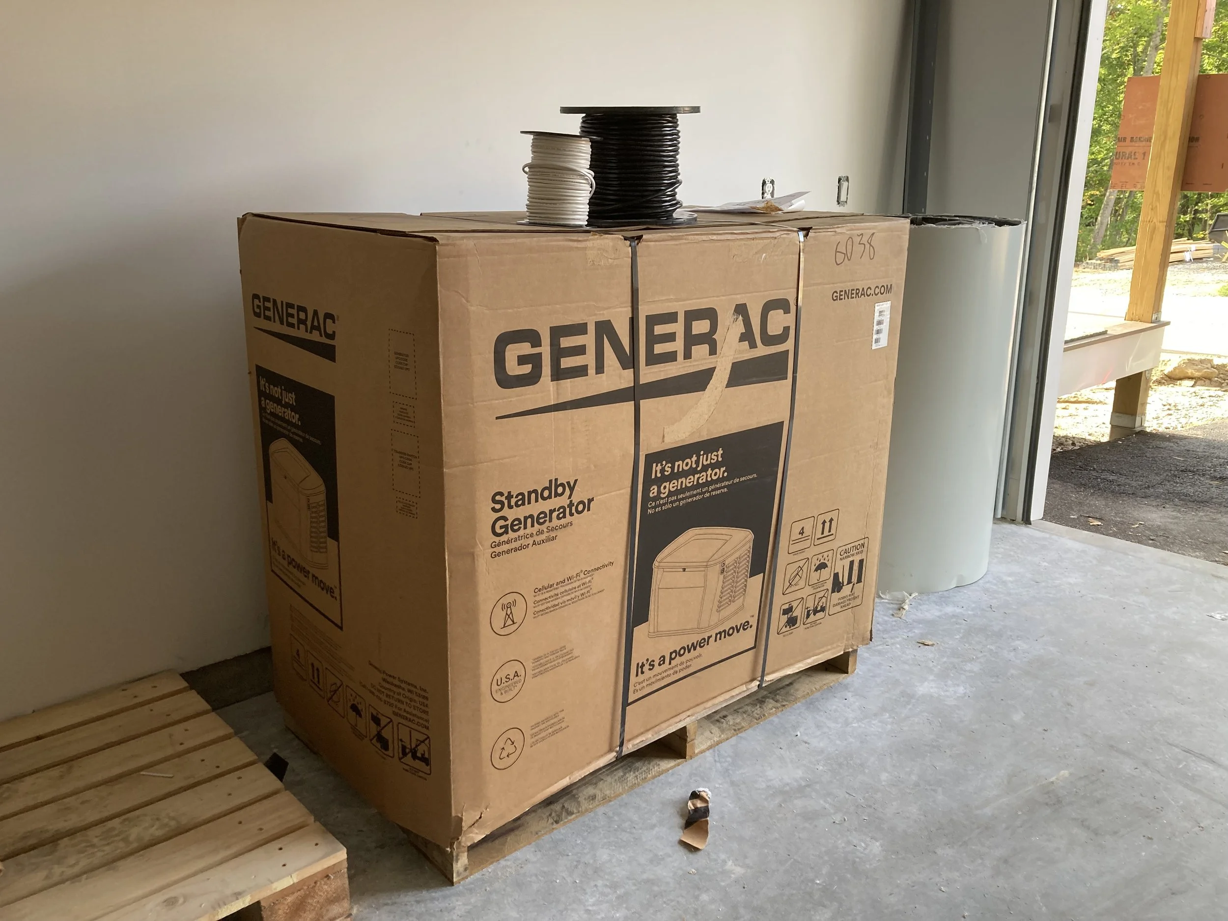 Large cardboard box labeled Generac standby generator, placed on a wooden pallet inside a garage. There are spools of wire on top of the box, and wooden boards to the side.