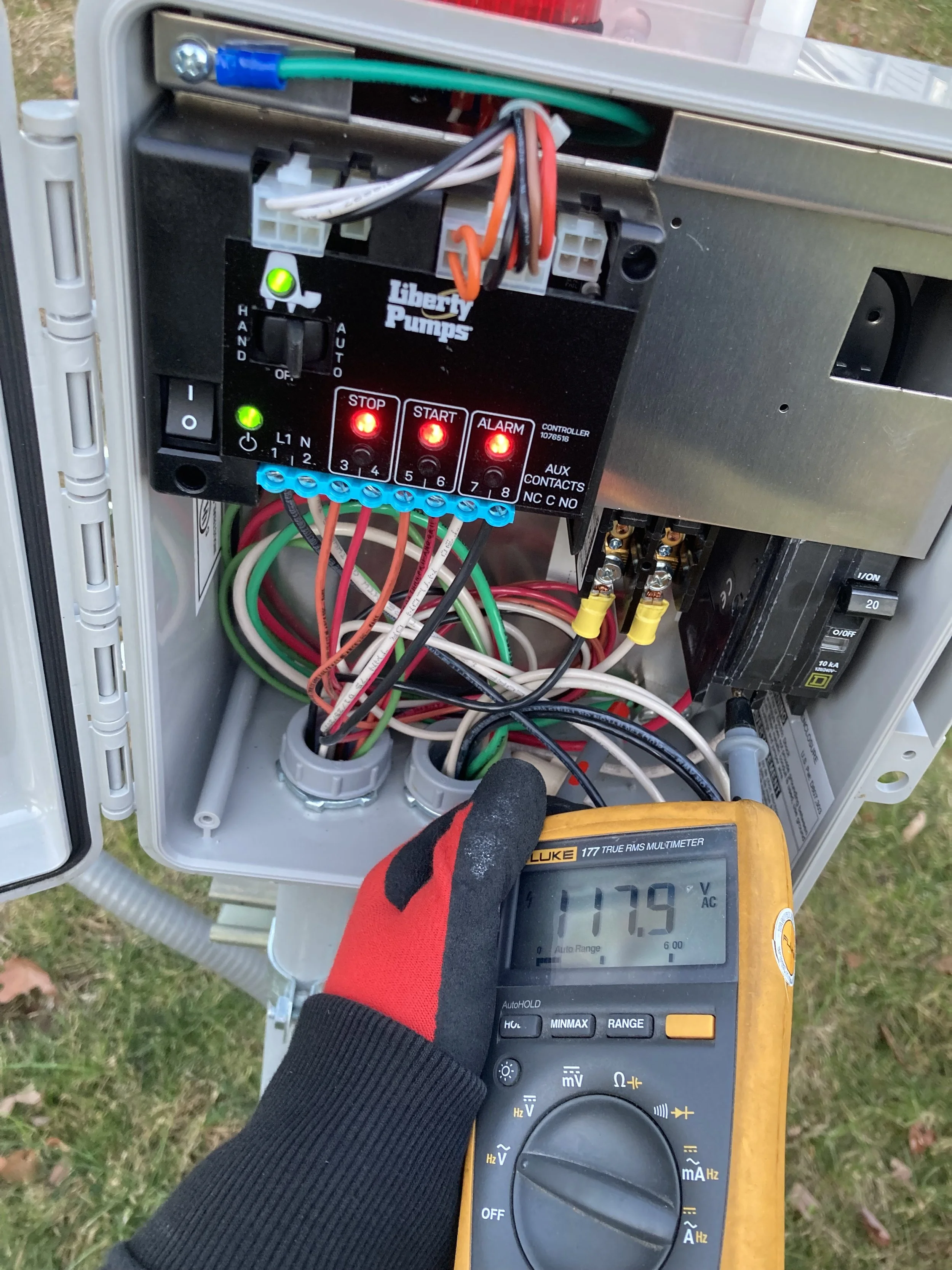 A person wearing a red and black glove is measuring 117 volts with a yellow digital multimeter connected to an electrical panel containing wires and a control unit.