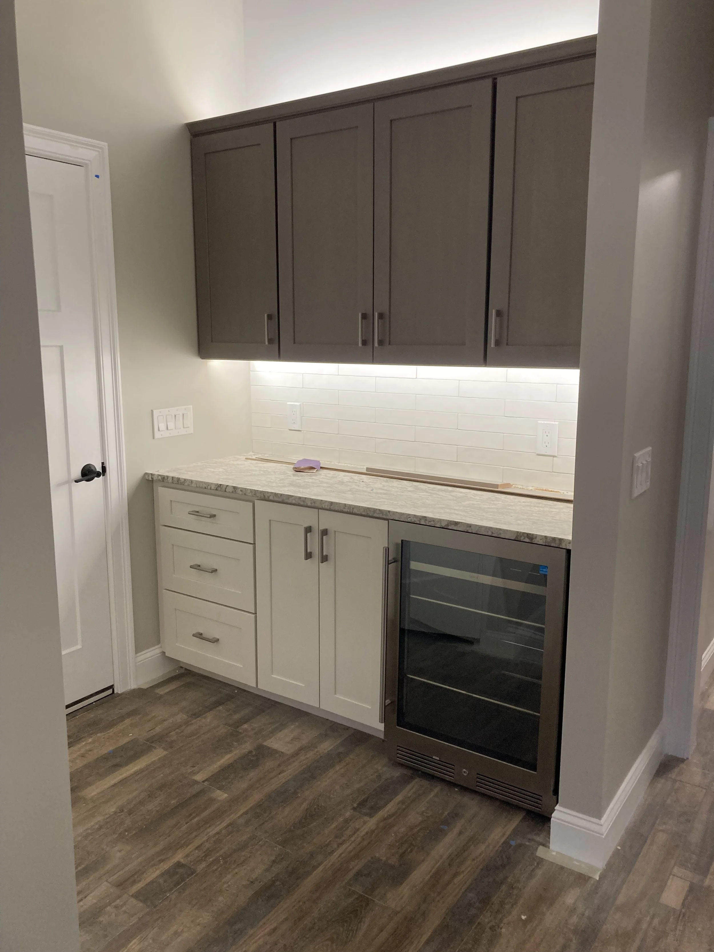 A small kitchen corner with beige and gray cabinetry, a granite countertop, a white brick tile backsplash, and a built-in wine refrigerator, with hardwood flooring and a white door.