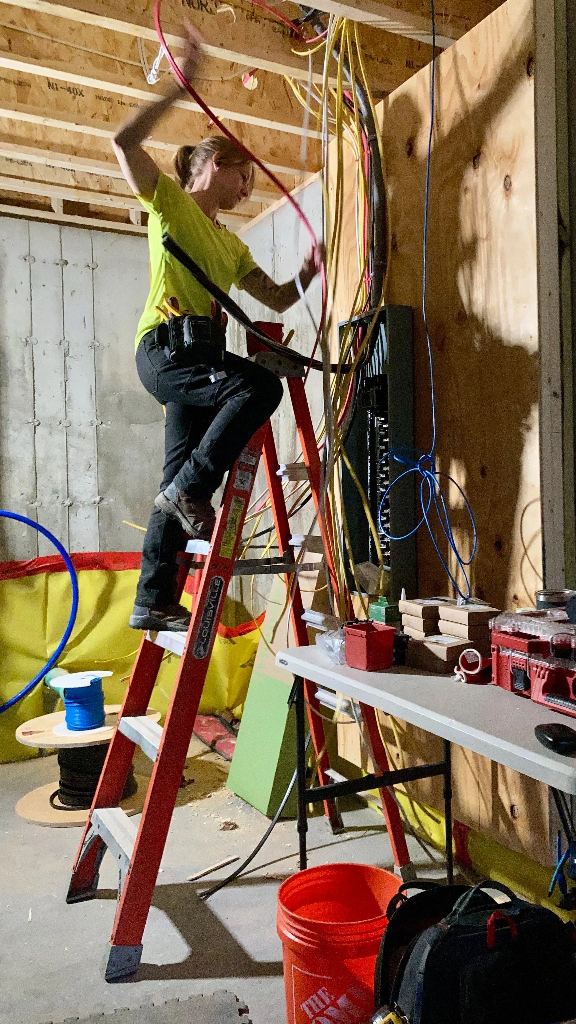 A woman working on electrical wiring inside a building under construction, standing on a red ladder with tools around her.