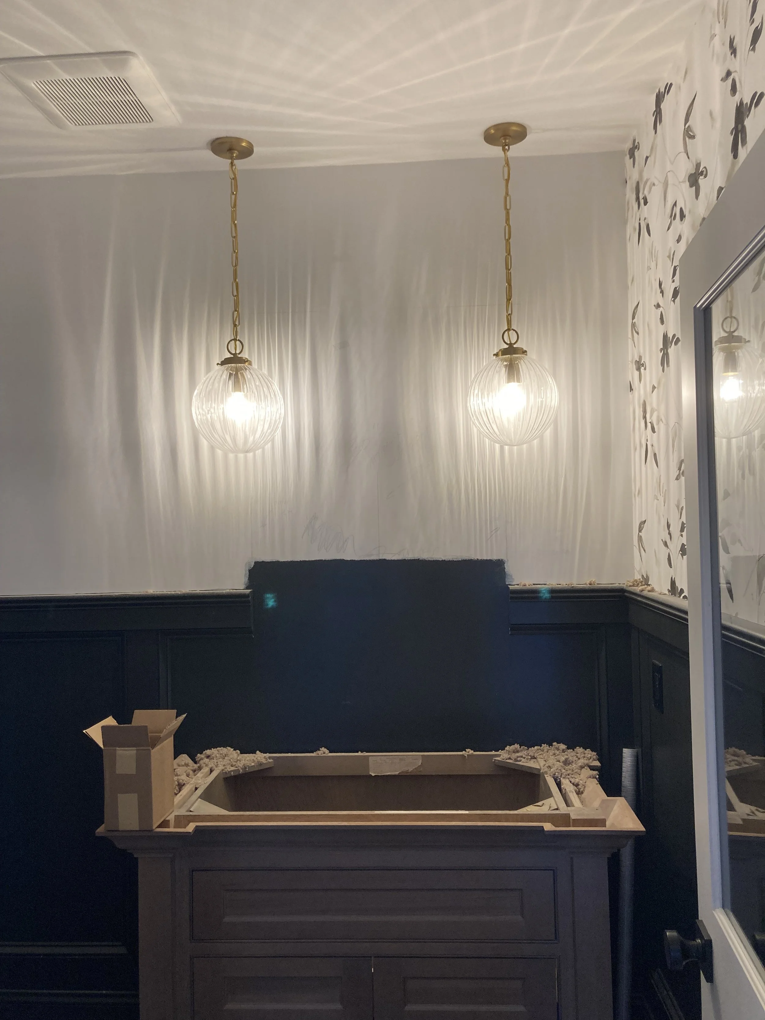 Two pendant lights with ribbed glass globes hanging from a white ceiling. A black wainscot runs along the wall below. A table is under construction, with a cardboard box and packing material on top.