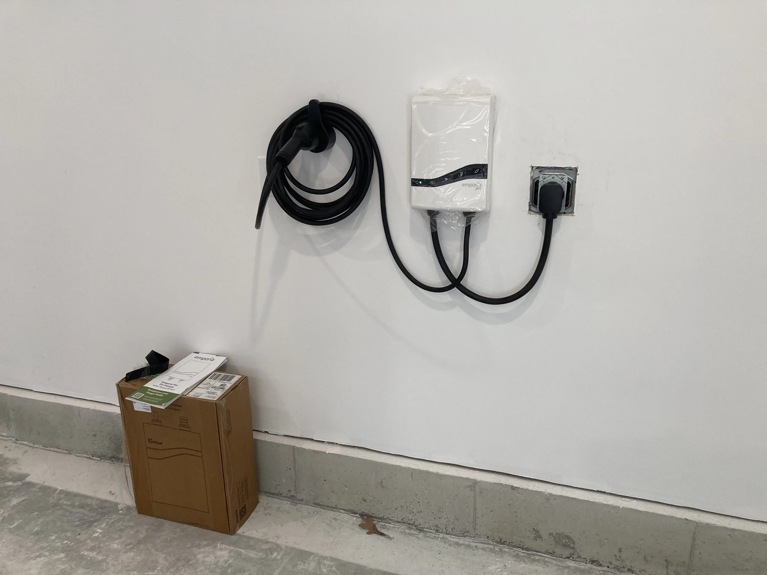 Electric vehicle charging station with a charging cable attached, mounted on a white wall next to an electrical outlet, and a cardboard box on the floor nearby.