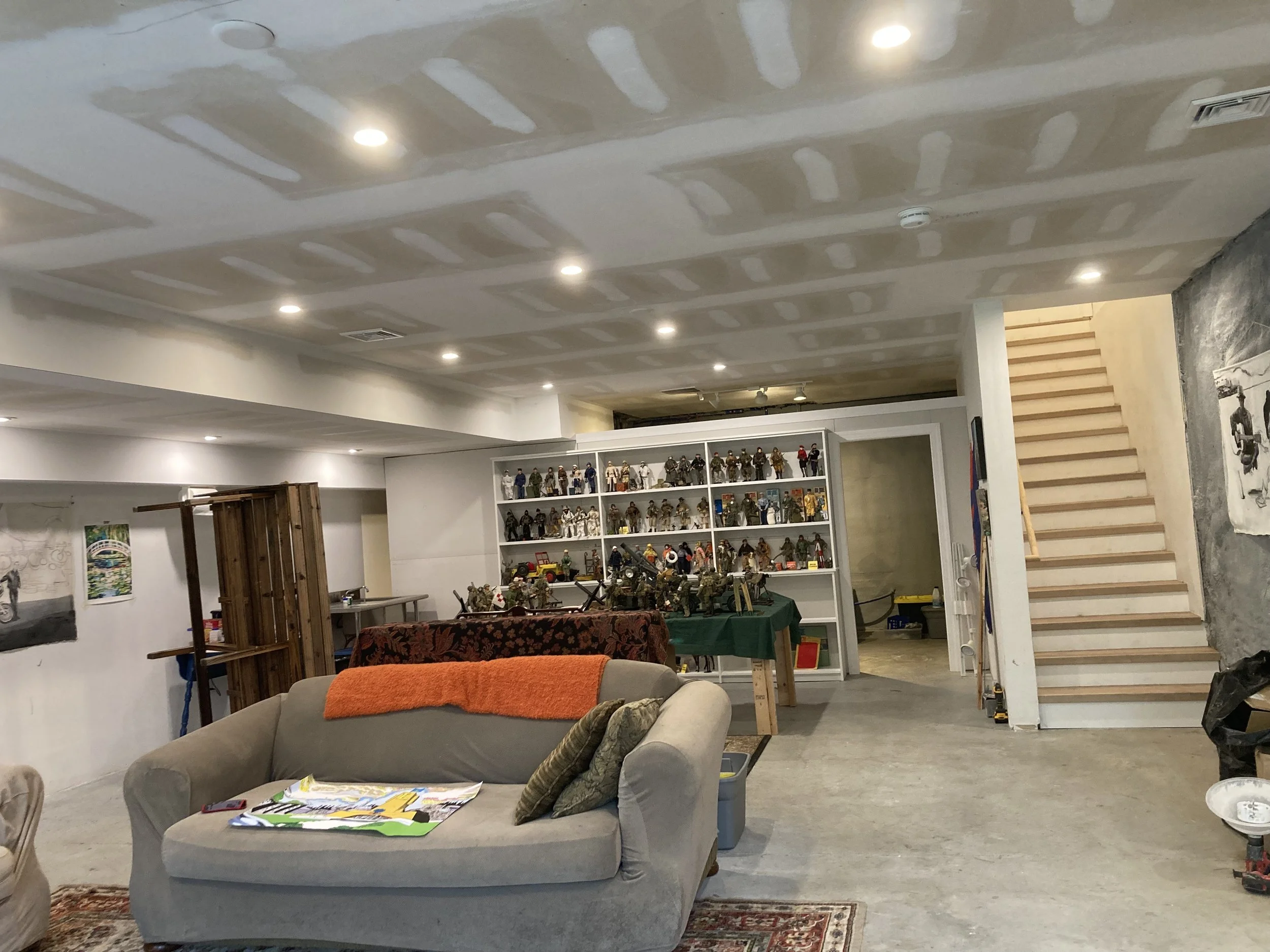 Living room with ceiling drywall, recessed lighting, stairs, a display shelf with toys, a beige couch with a rust-colored blanket, and a small table with magazines and pillows.