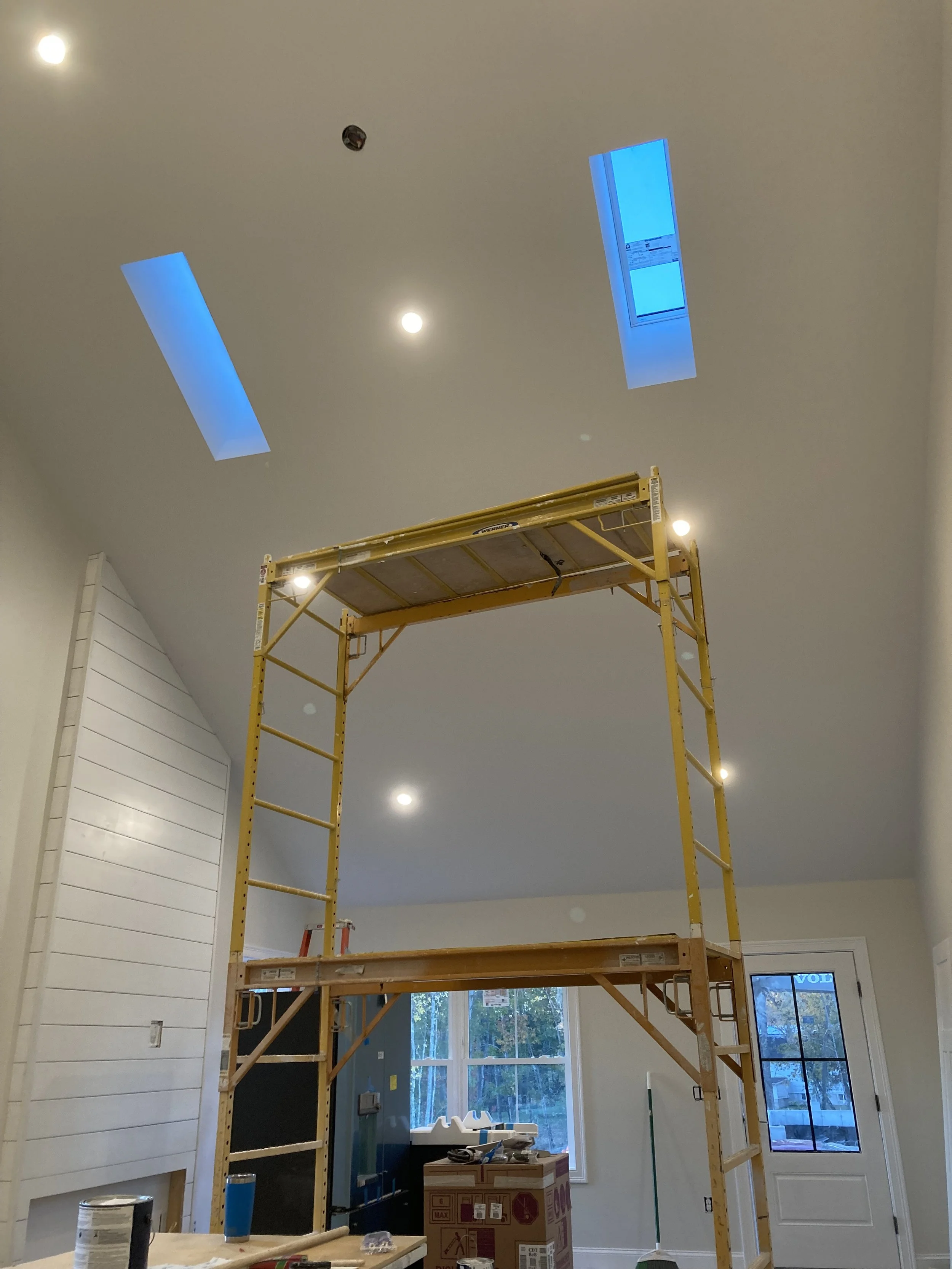 Room under construction with a yellow scaffold in the center, white shiplap on the wall, large window, and ceiling with recessed lights and skylights.