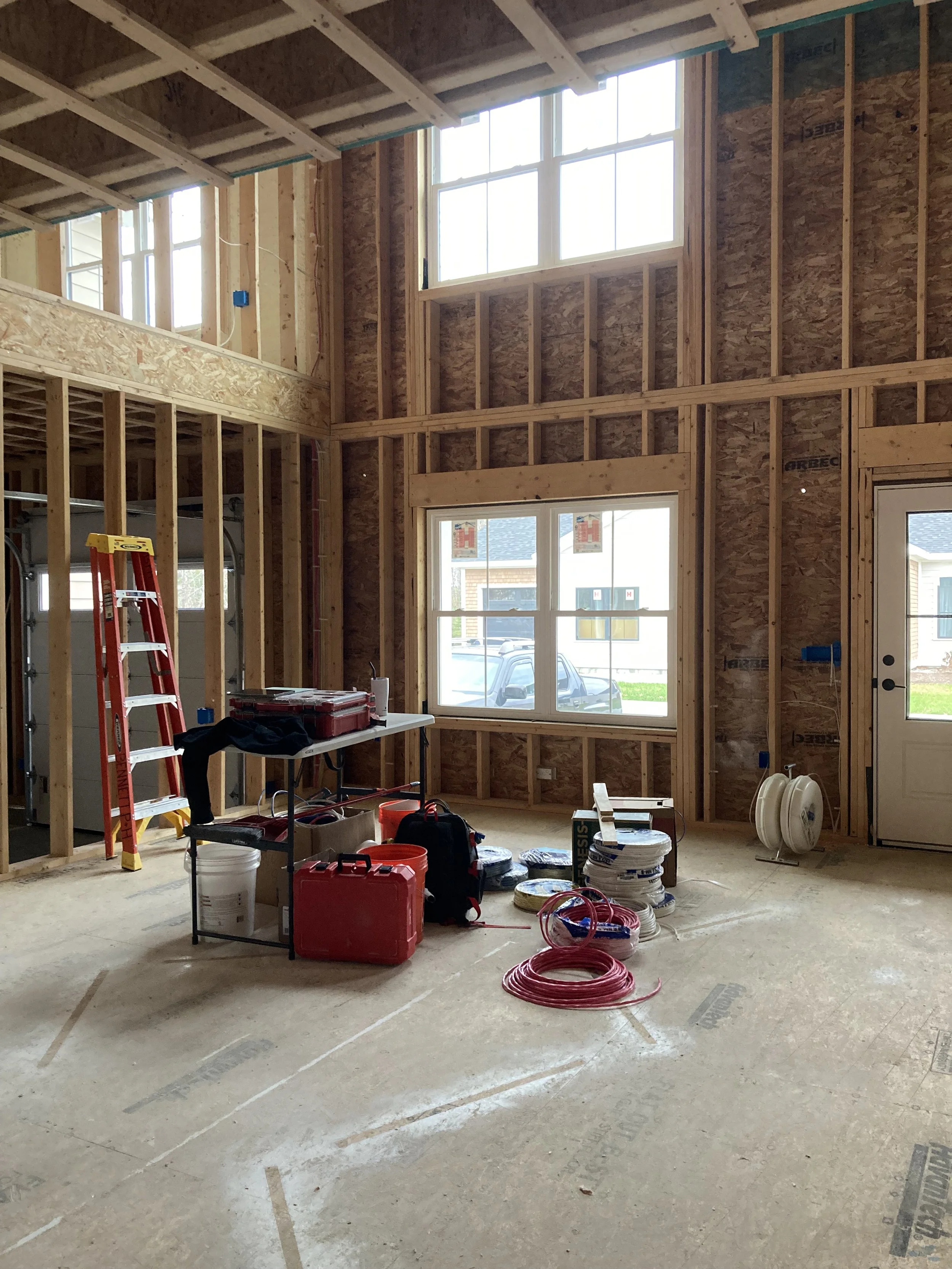 Interior view of a house under construction with exposed wooden framing, windows, and construction tools and materials on the floor.