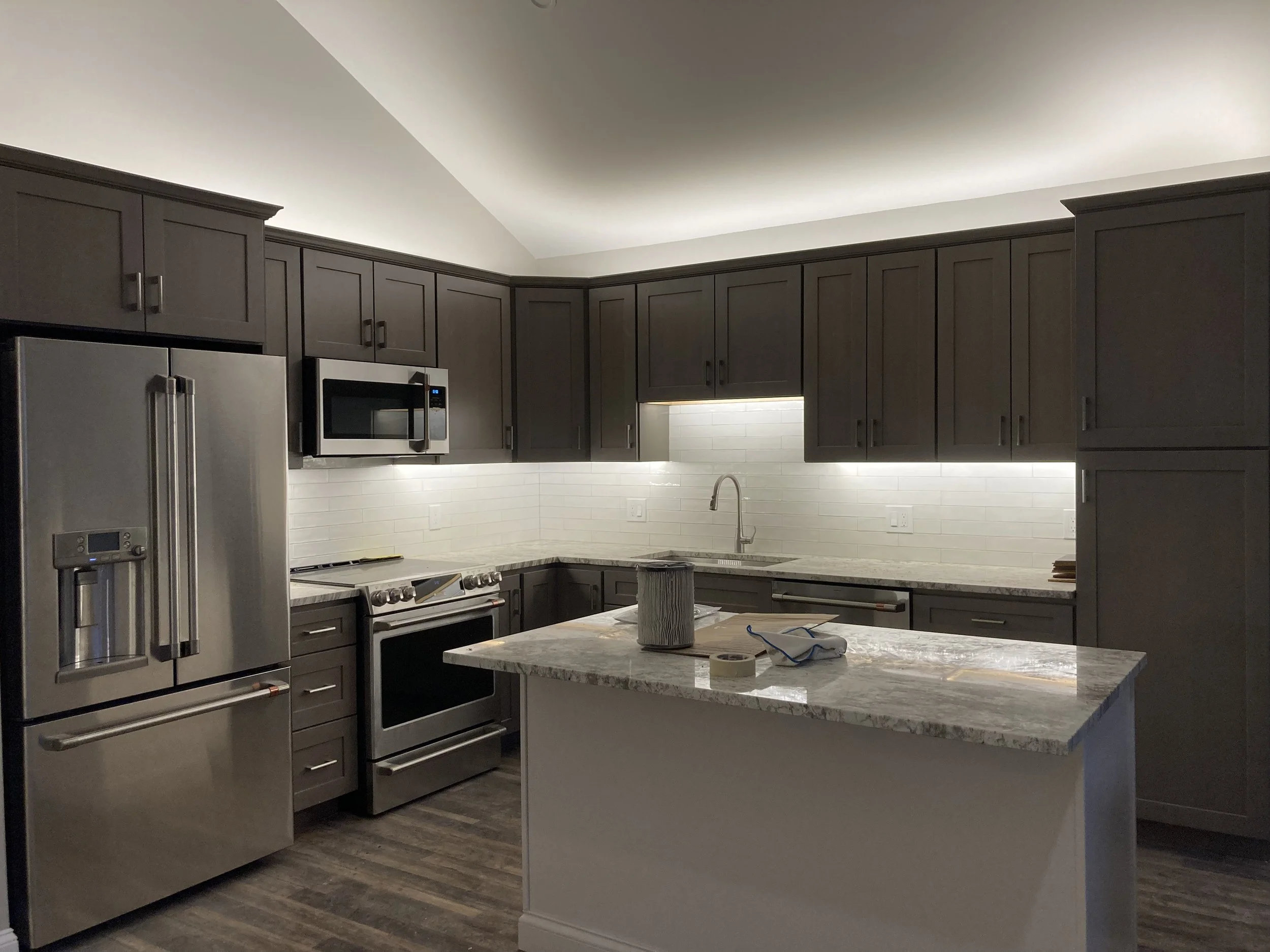Modern kitchen with gray cabinets, stainless steel appliances, marble countertop island, and white tile backsplash, under warm overhead lighting.