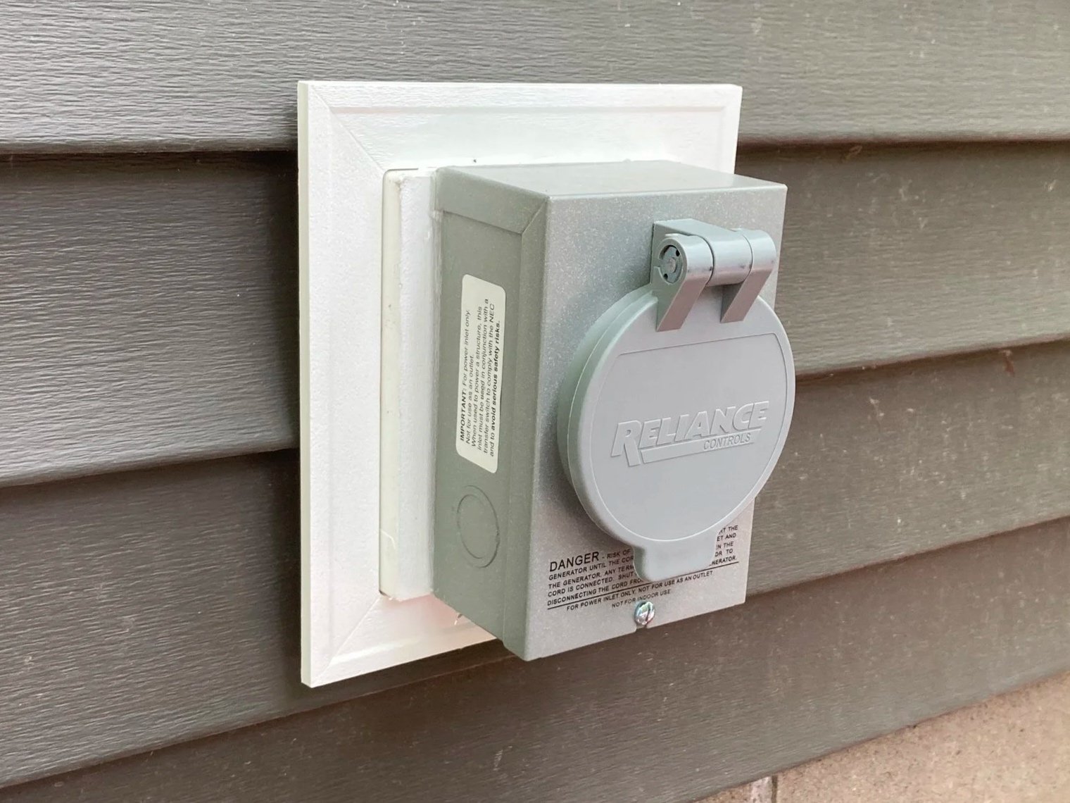 An outdoor electrical generator with a cover labeled 'Reliance Controls' mounted on a house wall.