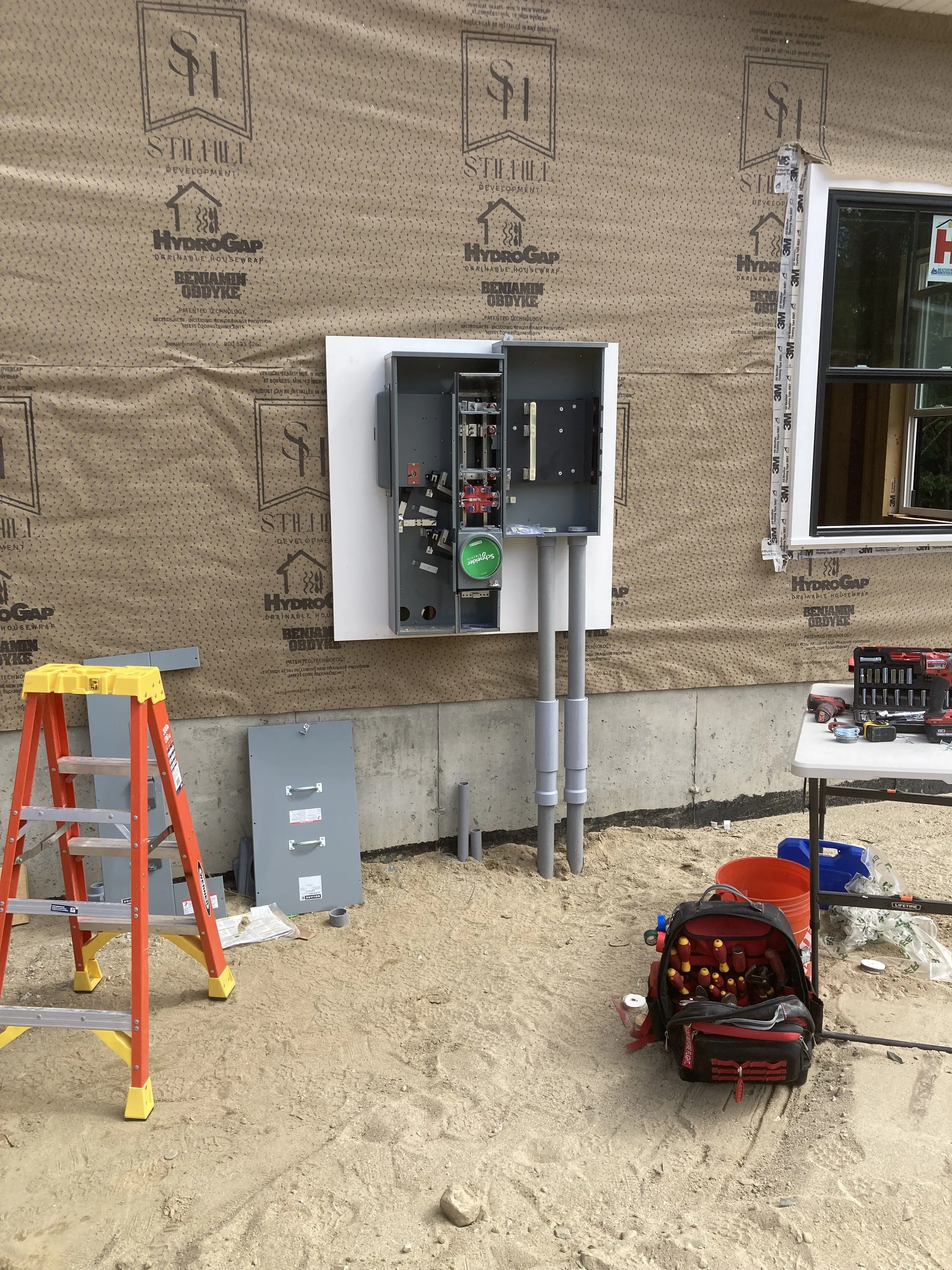 Electrical meter box being installed on the exterior of a house under construction, with tools, ladder, and construction materials around.