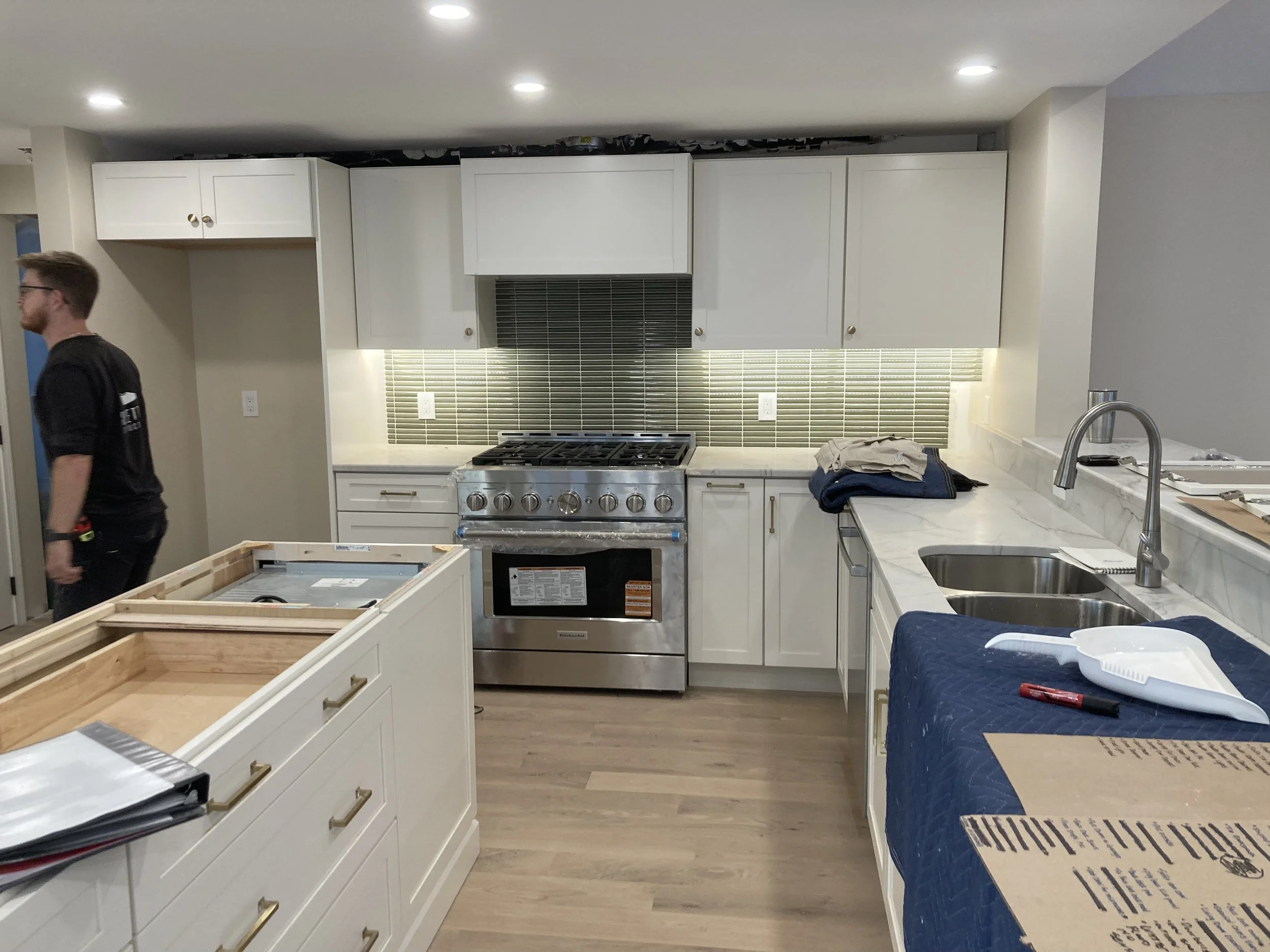 Kitchen with white cabinets, stainless steel stove, gray backsplash, marble countertops, and a double sink. A person is walking past on the left, and there are some items and boxes on the counters and island.