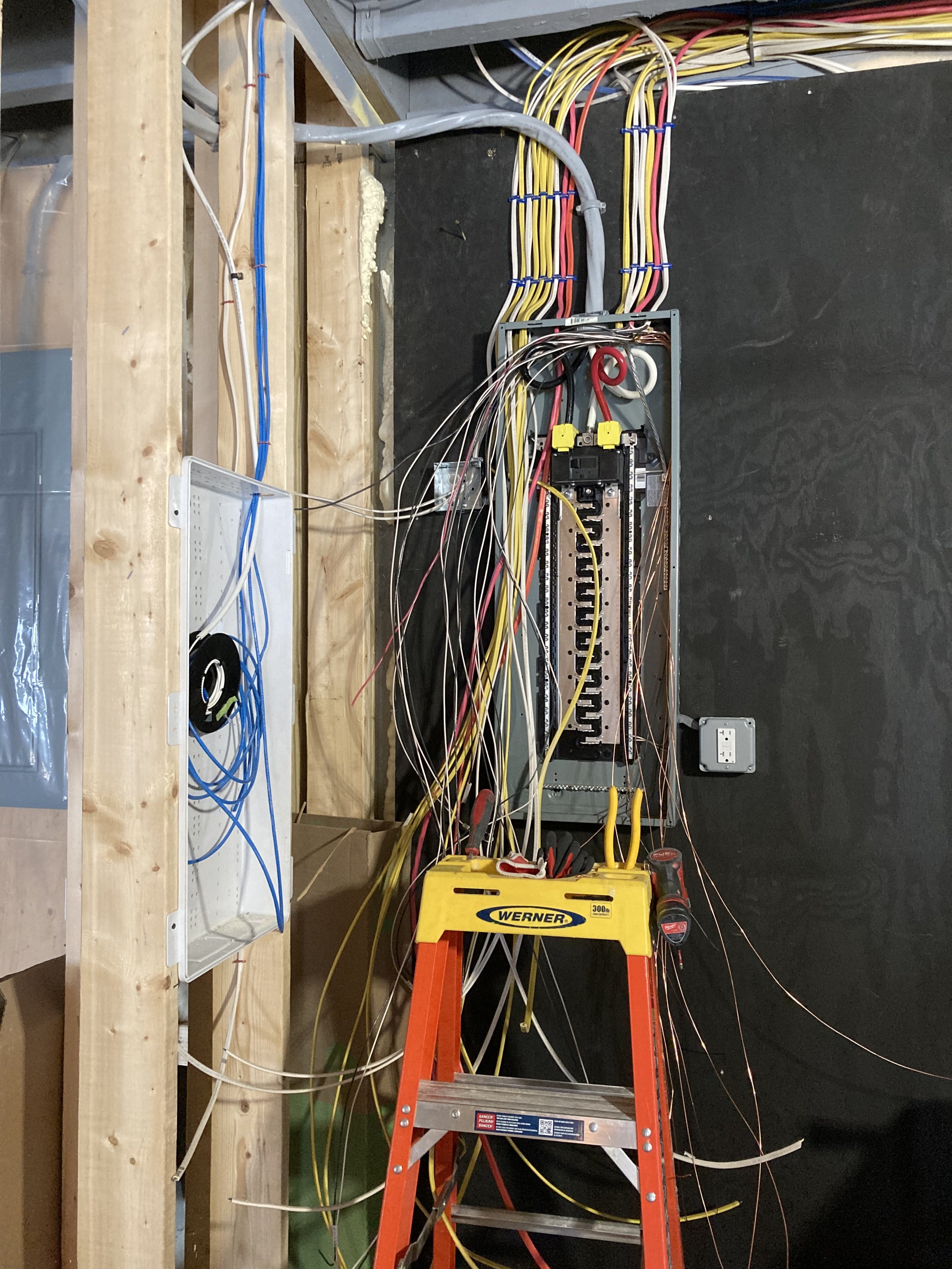 Electrical wiring and components mounted on a black wall, with an orange ladder in front, and a wooden framework on the left side.