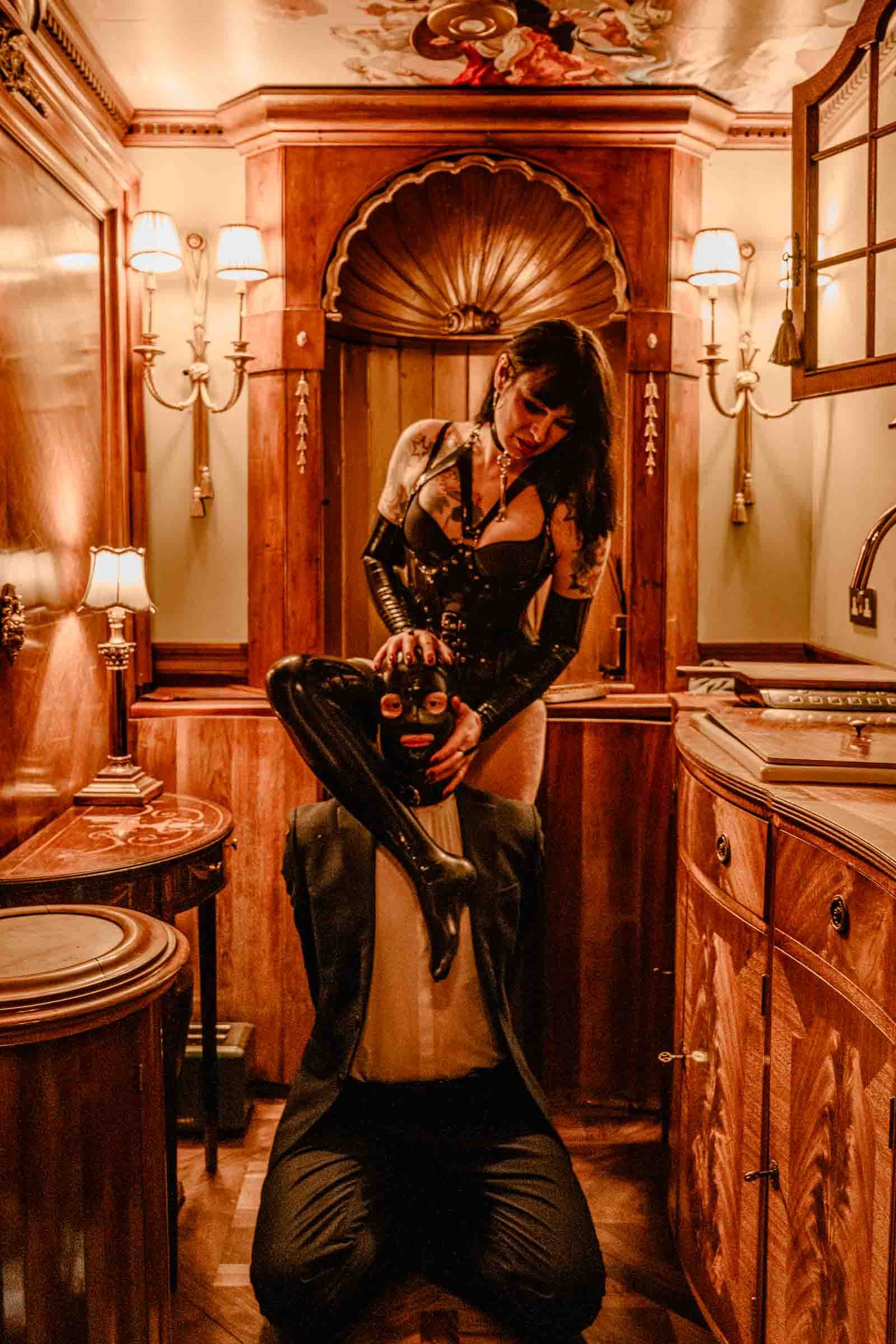 A woman with black hair and tattoos wearing black latex clothing and gloves, sitting on a man in black clothing and a mask, in a warmly lit, vintage-style wooden room with ornate wall sconces and a painted ceiling.
