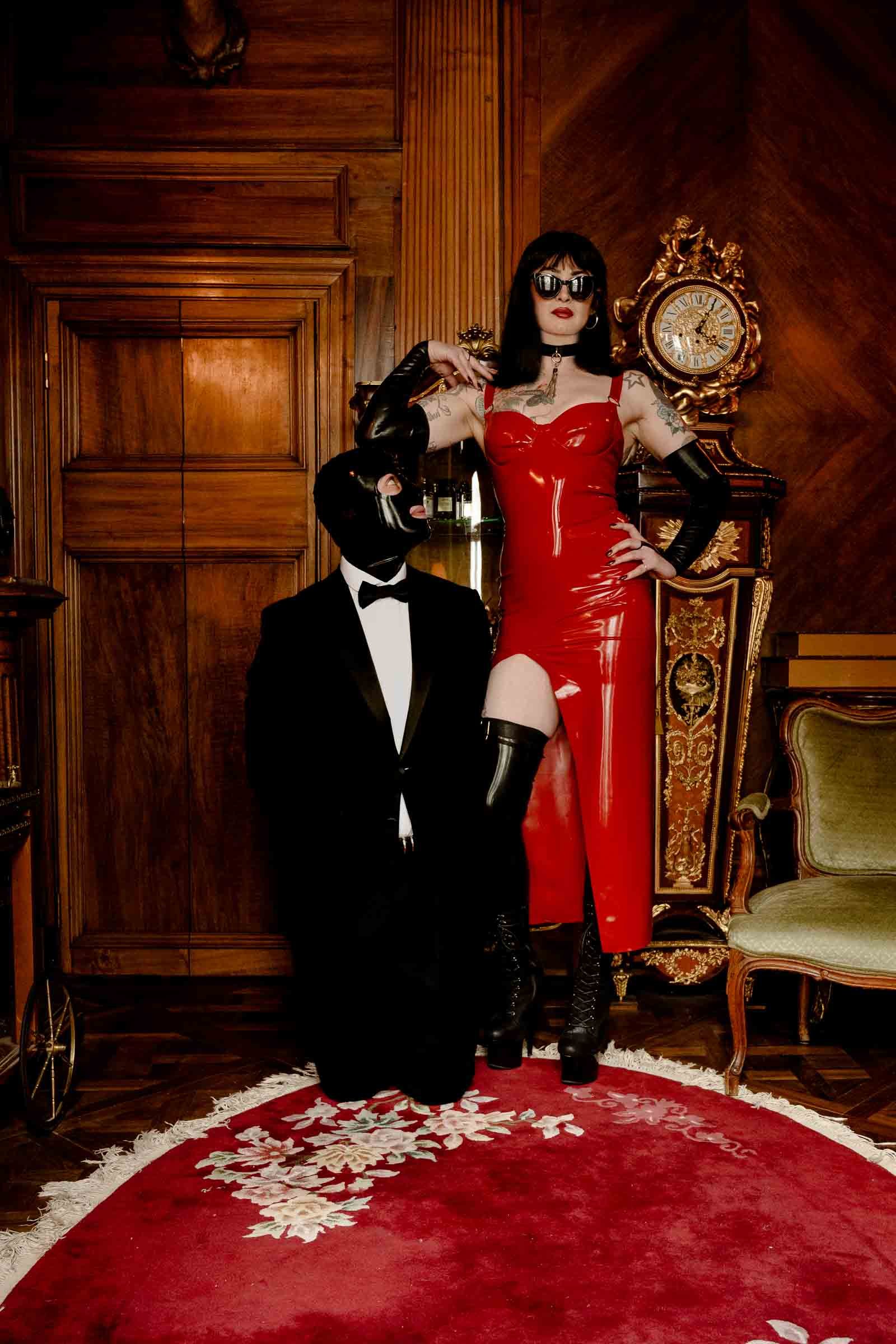 A woman dressed in a red latex gown with high thigh slit, black gloves, and sunglasses, standing inside a lavish room with wooden paneling, an ornate clock, and antique furniture. She is accompanied by a person in a black tuxedo and mask kneeling on 