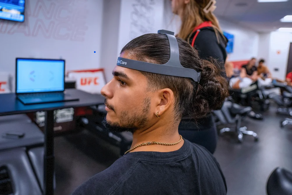 Young man seated in a gym wearing a head-mounted virtual reality device with long hair tied in a bun, surrounded by other people and gym equipment.