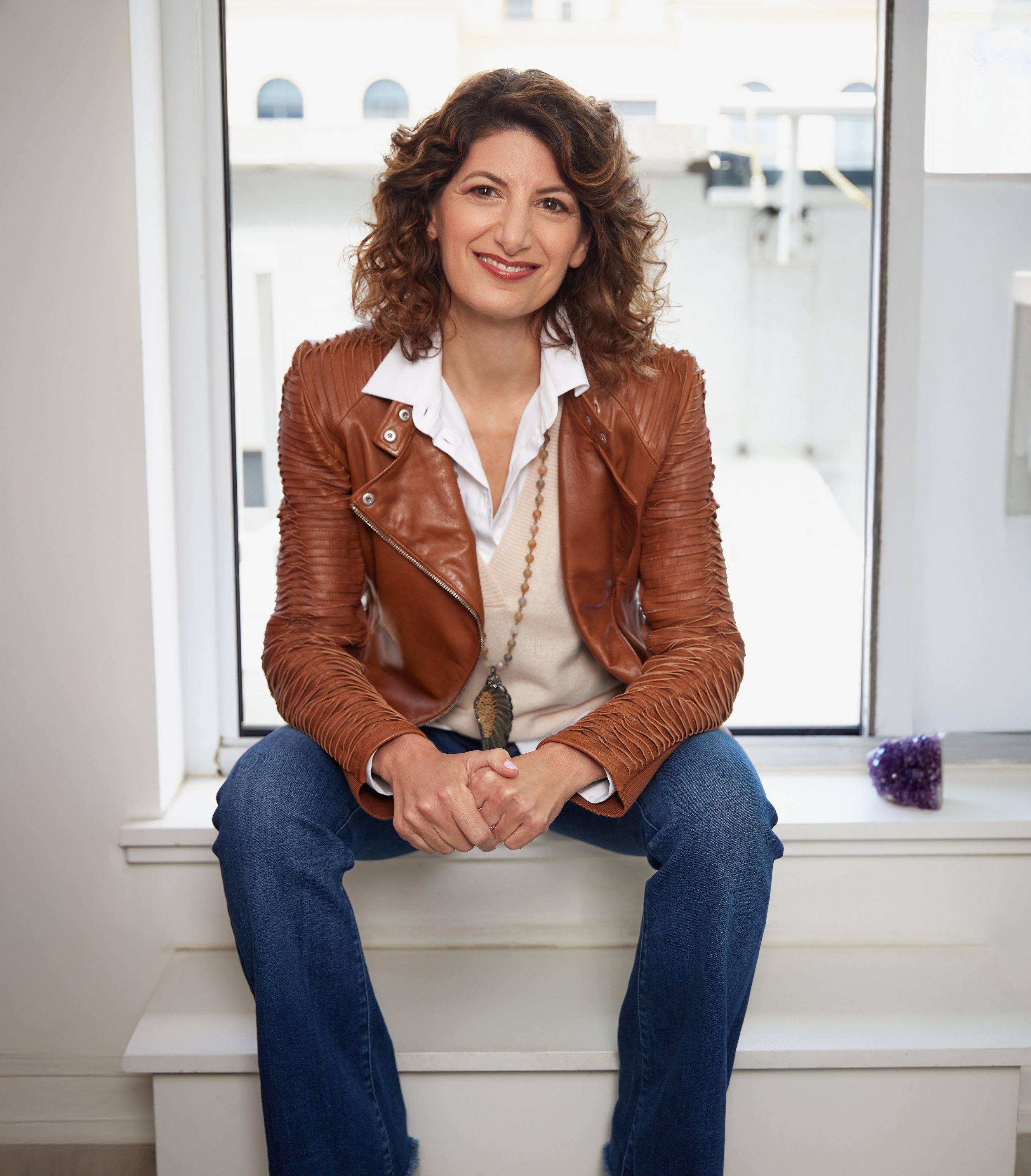 Stacey Tessis wearing a brown jacket and jeans, sitting smiling in a windowsill.