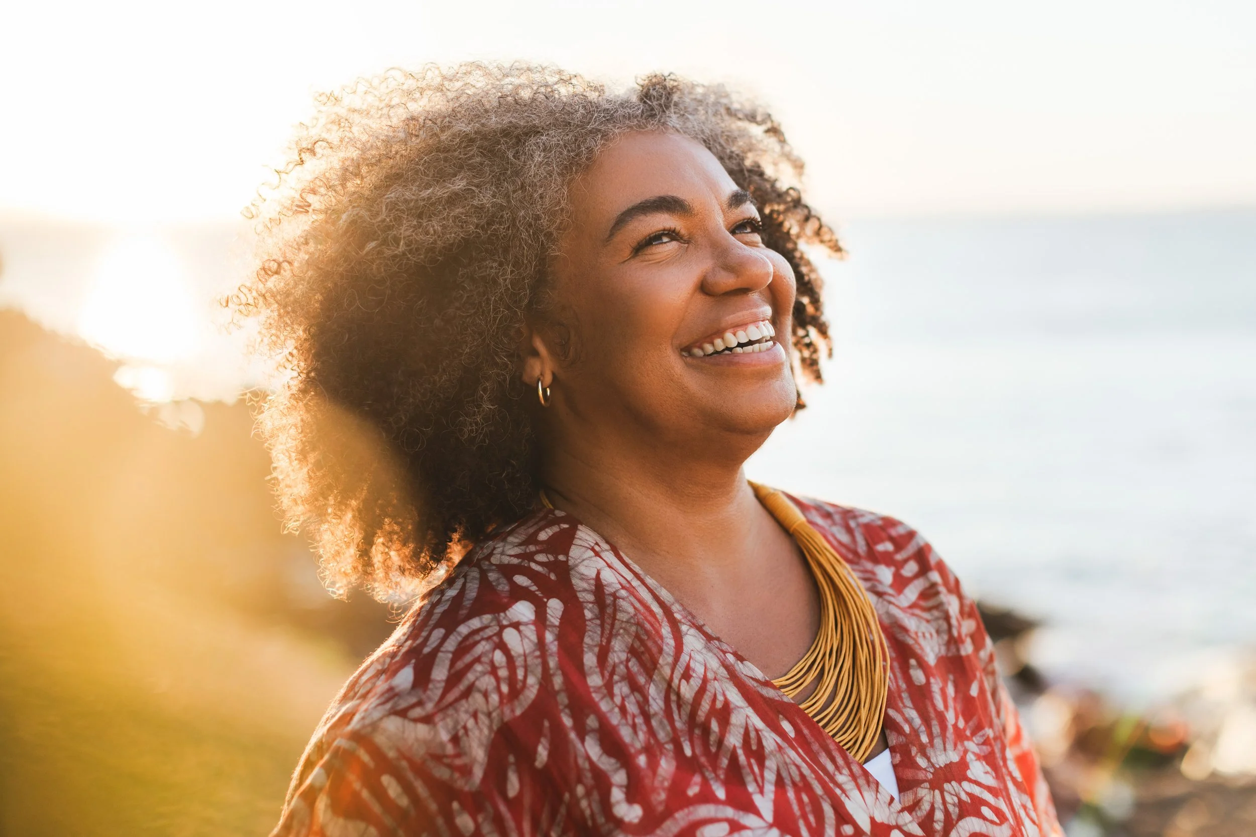 Woman with tight curly hair smiling and looking off into the distance outside.