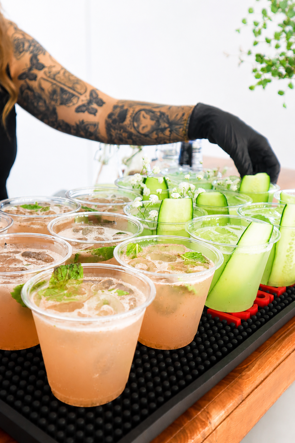 A person with a tattooed arm wearing a black glove is arranging glasses of pink citrus drinks garnished with fresh green herbs on a bar or table, with cucumber slices and small white flowers as decoration.