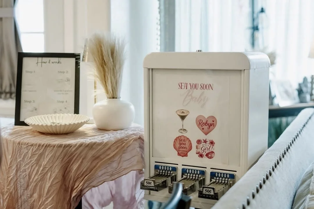 A white box with baby shower-themed stickers and raffle tickets in front, placed on a table covered with pink fabric, next to a white vase with pampas grass, and a picture frame in the background.