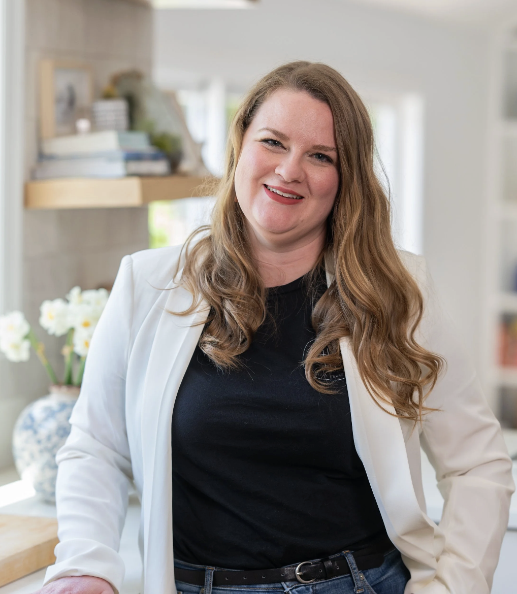A woman with long, wavy, auburn hair wearing a white blazer and black shirt, smiling in a bright kitchen with white flowers in a vase in the background.