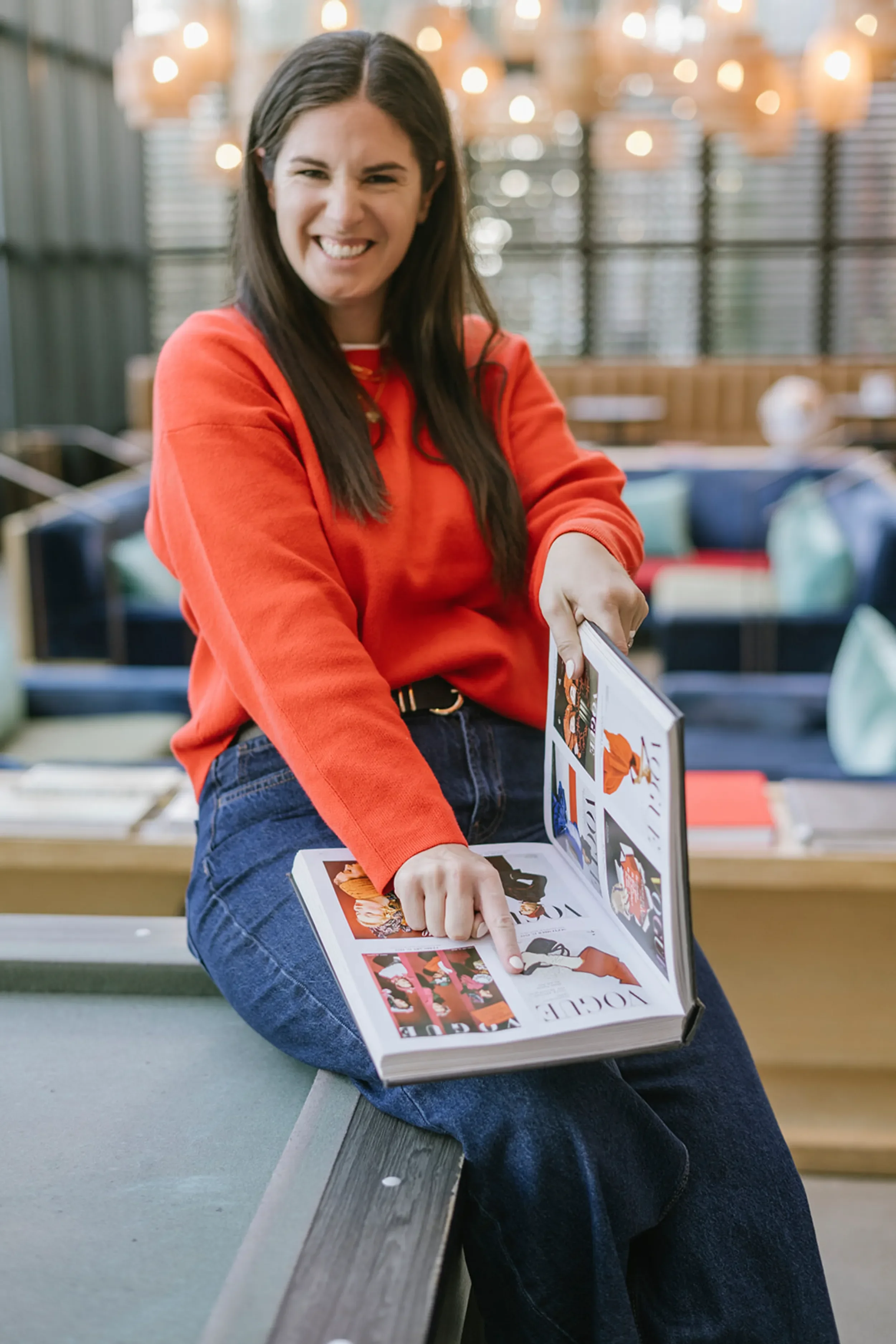 A young woman with long dark hair, wearing a red sweater and blue jeans, sitting on a bench in a bright, modern space, smiling and pointing at a photo book with fashion images.